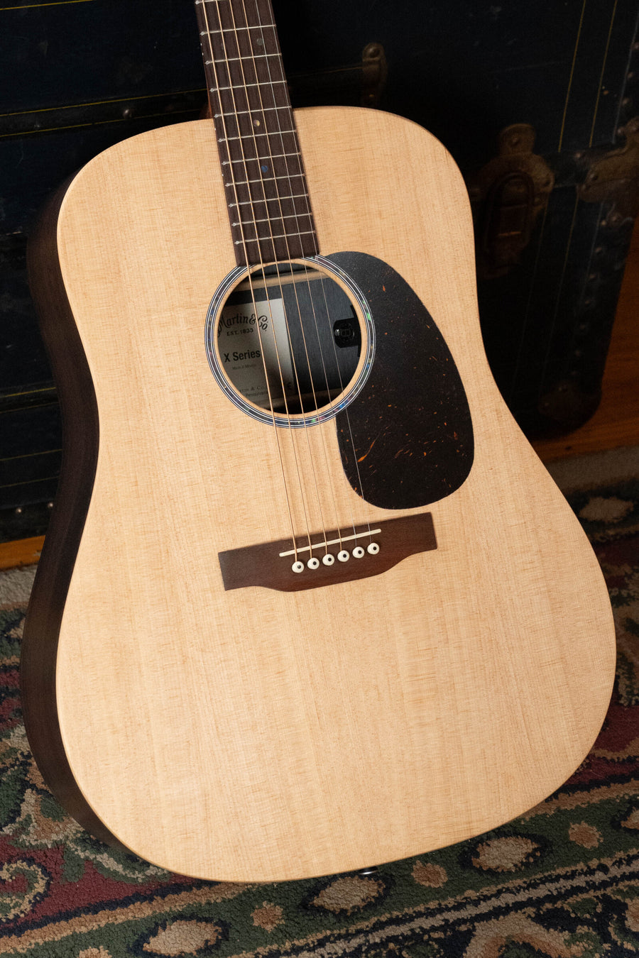 A close-up of the Martin & Co. D-X2E Sitka/HPL Brazilian Rosewood Dreadnought Guitar w/Martin E-1 Pickup #7868, featuring a light wood body, dark fretboard, and black pickguard, resting on a patterned rug before a dark trunk.