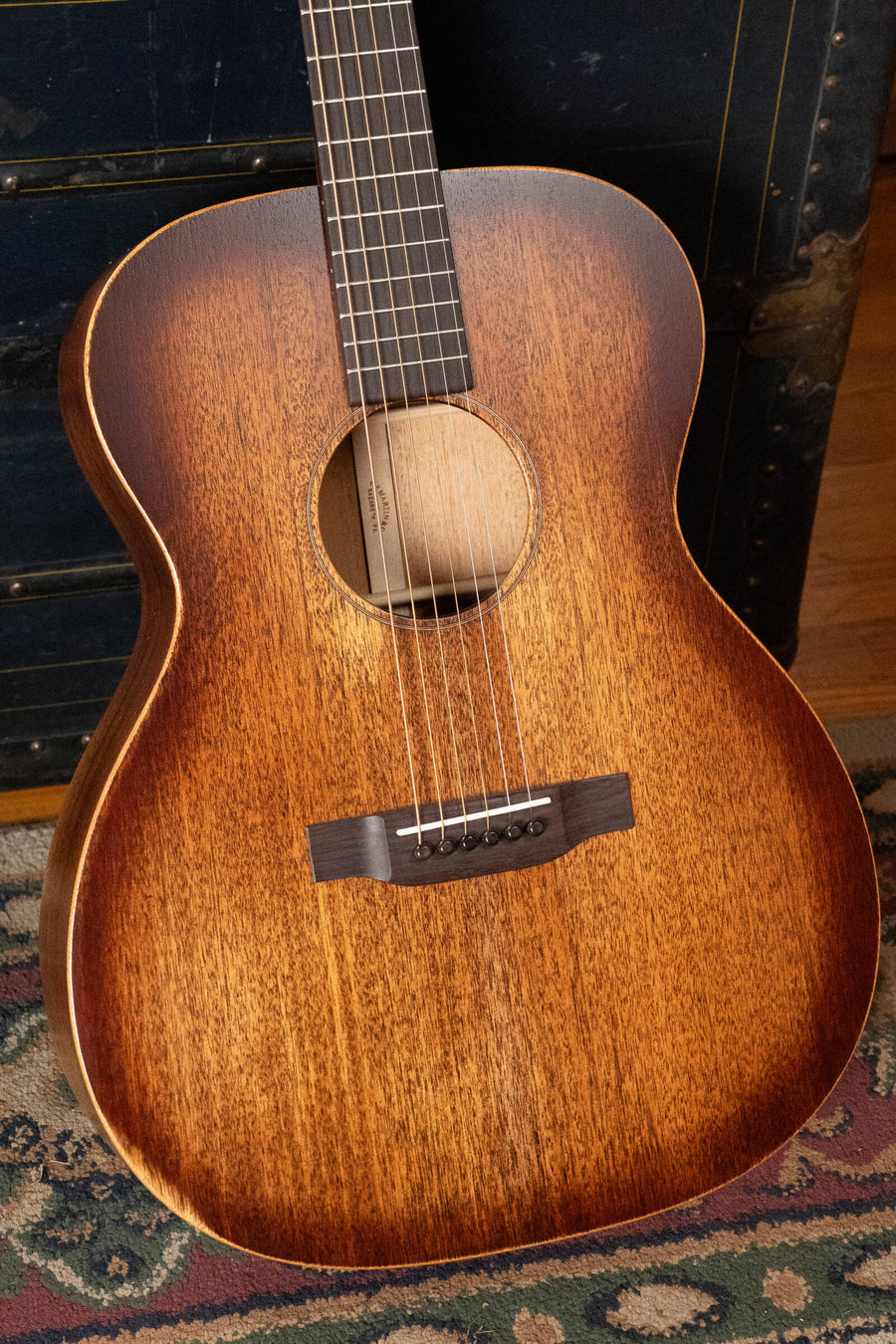A Martin & Co. OOO-15M StreetMaster All Mahogany OOO Acoustic Guitar #4910 stands upright on a patterned rug, showing its warm, natural wood finish, with part of a dark vintage trunk in the background.