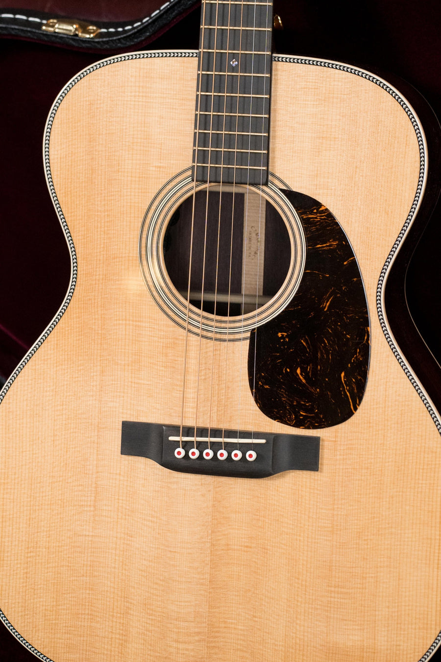 A close-up of the Martin & Co. OOO-28E Modern Deluxe Spruce/Indian Rosewood Guitar w/Electronics (#6398), showing its light solid spruce top, black marbled pickguard, bridge, strings, neck, and resting in a dark plush-lined case.