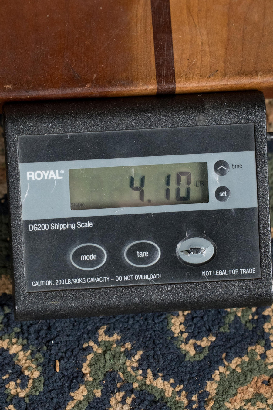 A digital shipping scale shows 4.10 lbs as it weighs a Martin & Co. OOO-28E Modern Deluxe Spruce/Indian Rosewood OOO Guitar w/Electronics #6398, resting on a patterned carpet next to wood, with mode, tare, and on/off buttons visible.