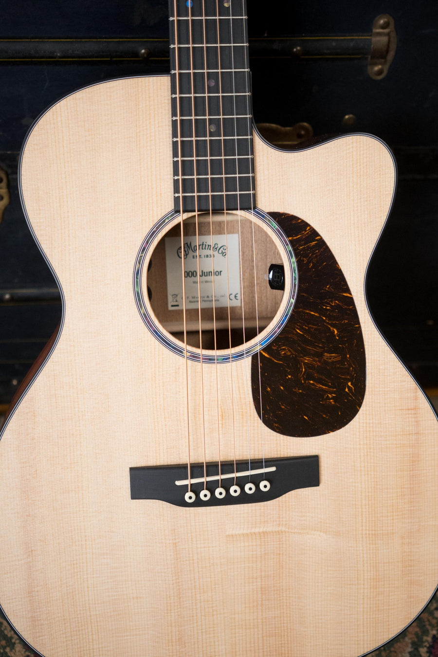 Close-up of a Martin & Co. OOO Jr CE Spruce/Sapele Cutaway Guitar (#4755), highlighting its sound hole, strings, and dark pickguard. The background is dark and out of focus. Built-in Martin electronics are featured.