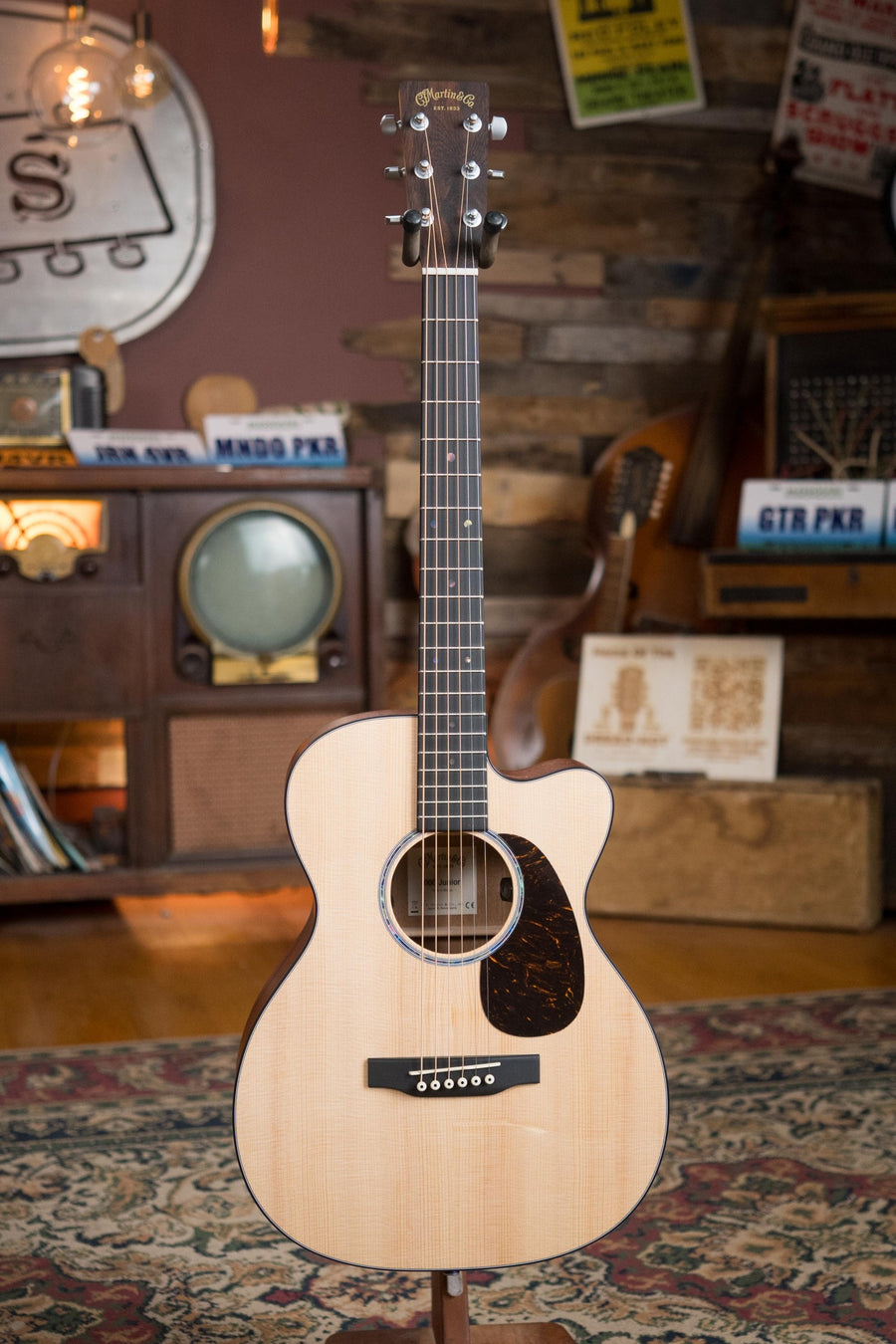 A Martin & Co. OOO Jr CE Spruce/Sapele Cutaway Guitar w/Martin Electronics (#4755) stands upright on a patterned rug in a cozy, vintage-themed room with a retro TV, wooden shelves, wall posters, and a dark pickguard.