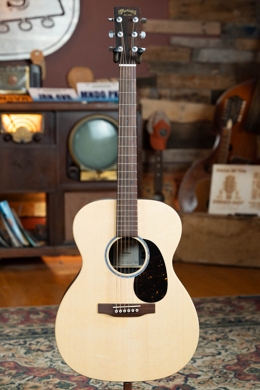 A Martin & Co. OOO-X2E Sitka/HPL Brazilian Rosewood guitar with E-1 Pickup #1554 and a dark pickguard stands upright in a cozy, vintage-style room accented by wood tones and musical instruments.