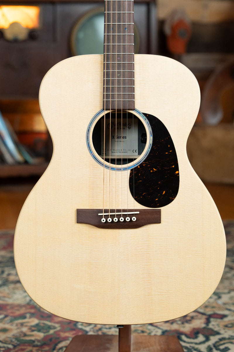 A close-up of the Martin & Co. OOO-X2E Sitka/HPL Brazilian Rosewood Guitar #1554 highlights its light wooden body, six strings, dark pickguard, and round soundhole, with a blurred vintage-themed background.