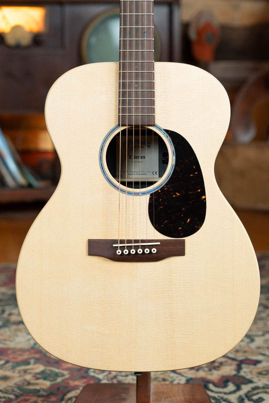 A close-up of the Martin & Co. OOO-X2E Sitka/HPL Brazilian Rosewood Guitar #1554 highlights its light wooden body, six strings, dark pickguard, and round soundhole, with a blurred vintage-themed background.