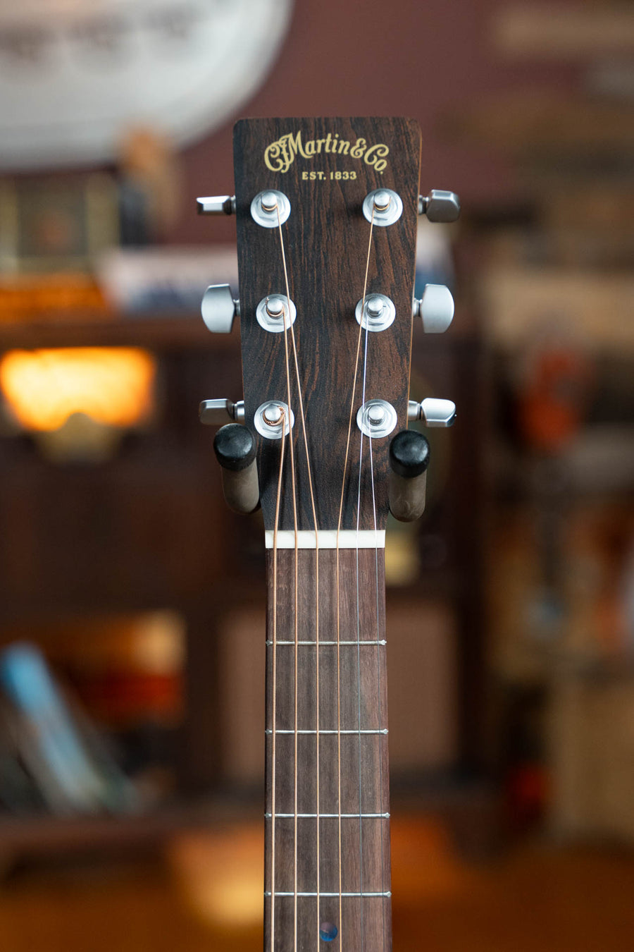 Close-up of the headstock and upper neck of a Martin & Co. OOO-X2E Sitka/HPL Brazilian Rosewood Guitar w/E-1 Pickup, showing tuning pegs, strings, and logo against a warmly lit, slightly blurred indoor background.