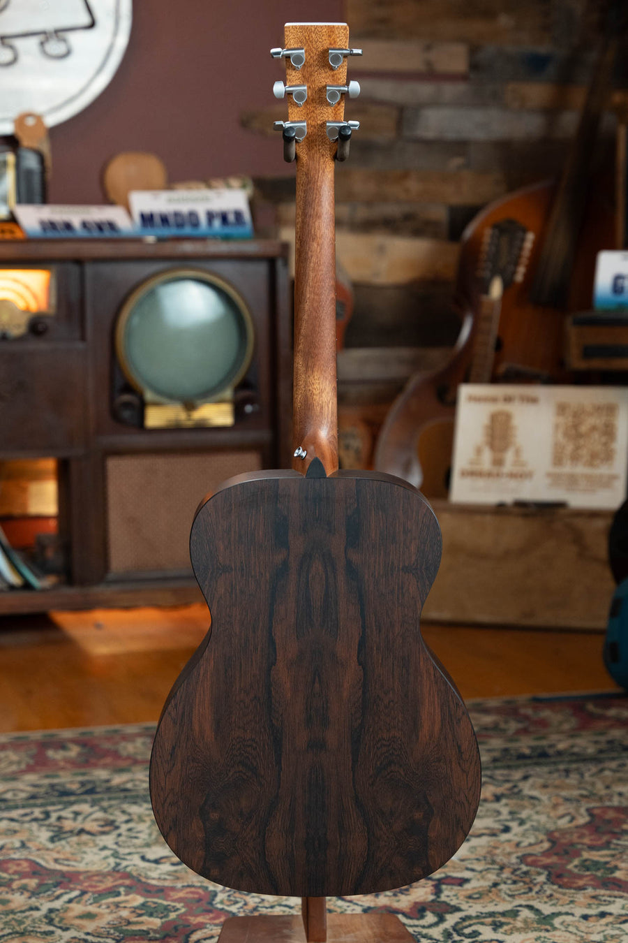 A Martin & Co. OOO-X2E Sitka/HPL Brazilian Rosewood OOO Guitar w/Martin E-1 Pickup #1554 is seen from the back, showcasing its dark Brazilian Rosewood finish in a cozy room with vintage decor and various musical instruments.