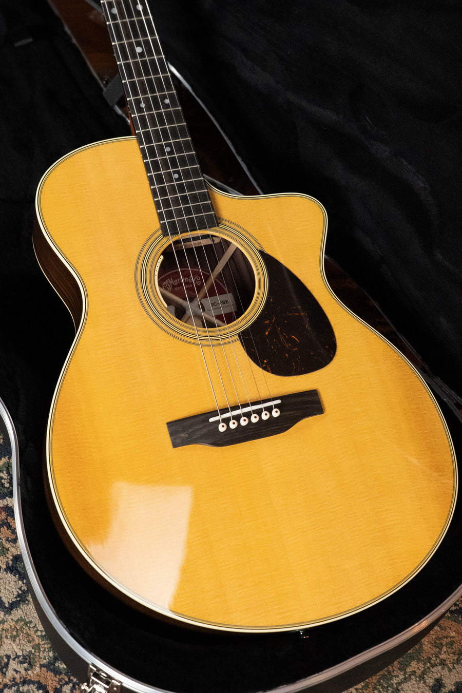 A close-up of the Martin & Co. SC-28E Spruce/Indian Rosewood S Body Cutaway Guitar with Fishman electronics, natural finish, black pickguard, and six strings, shown in an open black hard case.