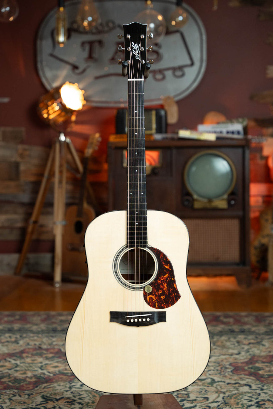 The Maton Guitars Master Built Adirondack/Red Cedar Dreadnought Guitar w/Electronics #0020 stands upright in a cozy, vintage-style room with warm lighting, wood walls, an old TV, and retro decor.