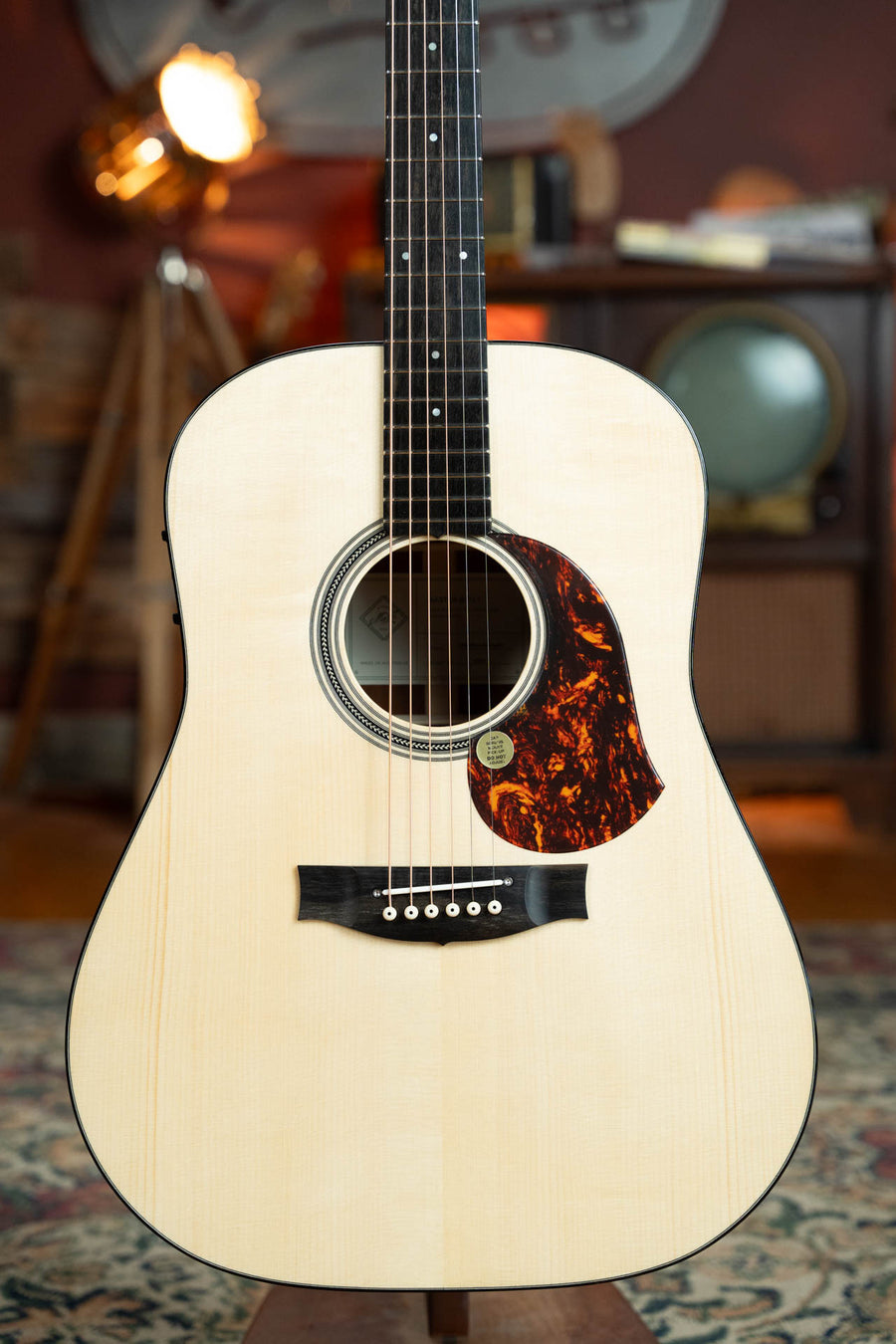 A close-up of the Maton Guitars Master Built Adirondack/Red Cedar Dreadnought Guitar w/Electronics (#0020) shows its detailed body, dark pickguard, and black fretboard, standing upright amid vintage decor and warm lighting.