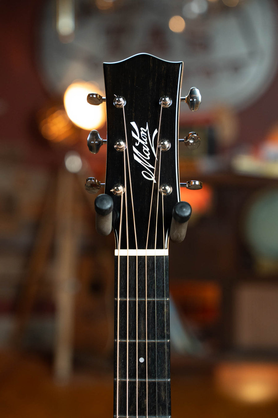 Close-up of a Maton Guitars headstock with chrome tuners and white script logo, set against a warmly lit, blurred background—capturing the elegance of the Maton Master Built Adirondack/Red Cedar Dreadnought w/Electronics #0020.