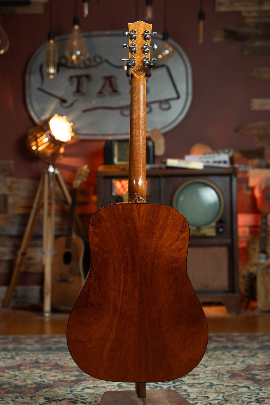 A Maton Guitars Master Built Adirondack/Red Cedar Dreadnought Guitar w/Electronics (#0020) stands upright with its back visible, set against vintage decor, a retro TV, and cozy warm lighting.