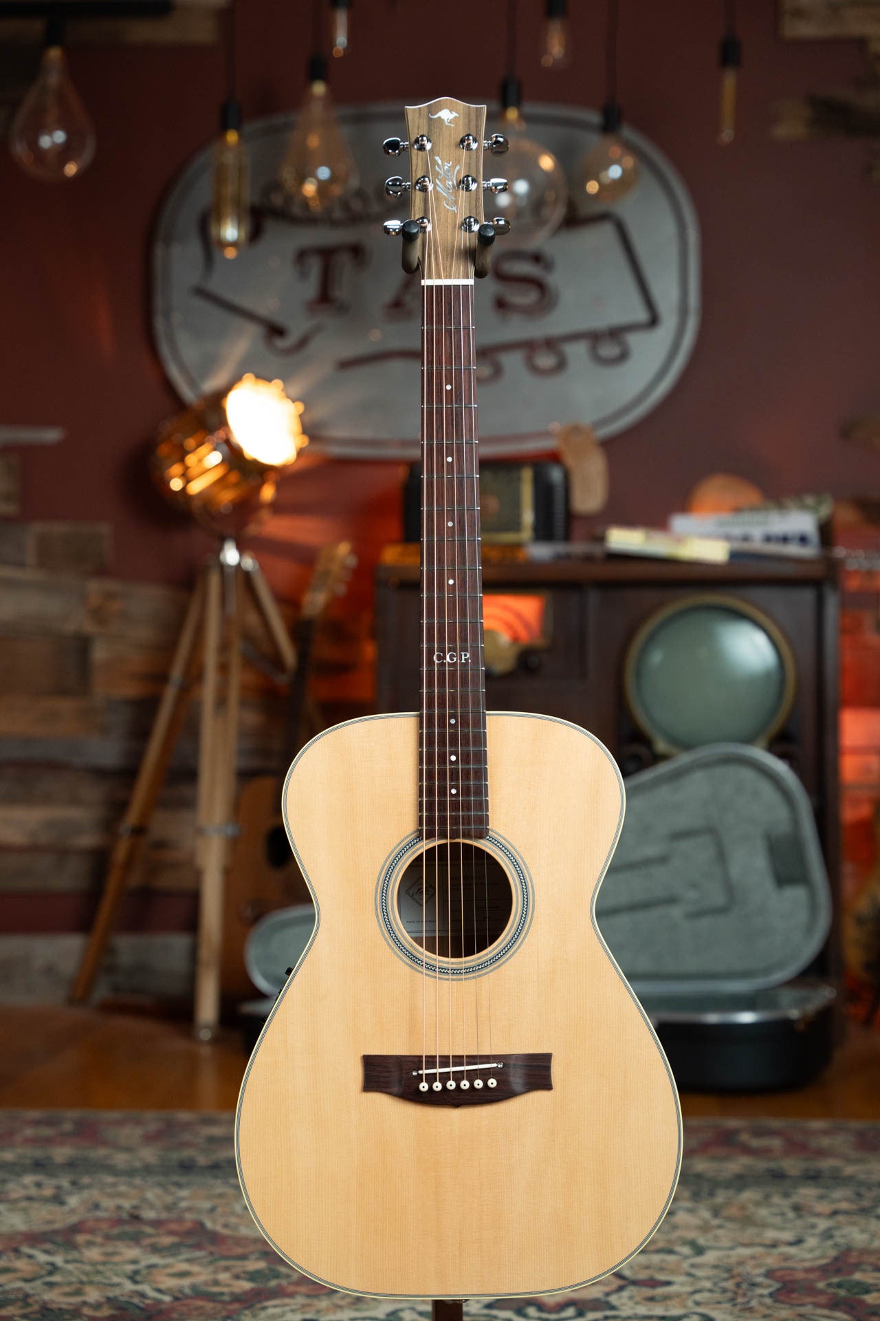 A Maton Guitars T.E. Personal Spruce/Queensland Maple Deep Body OO Guitar with electronics (#0085) stands upright in a cozy, vintage-style room filled with warm lighting, musical equipment, and decorative elements.