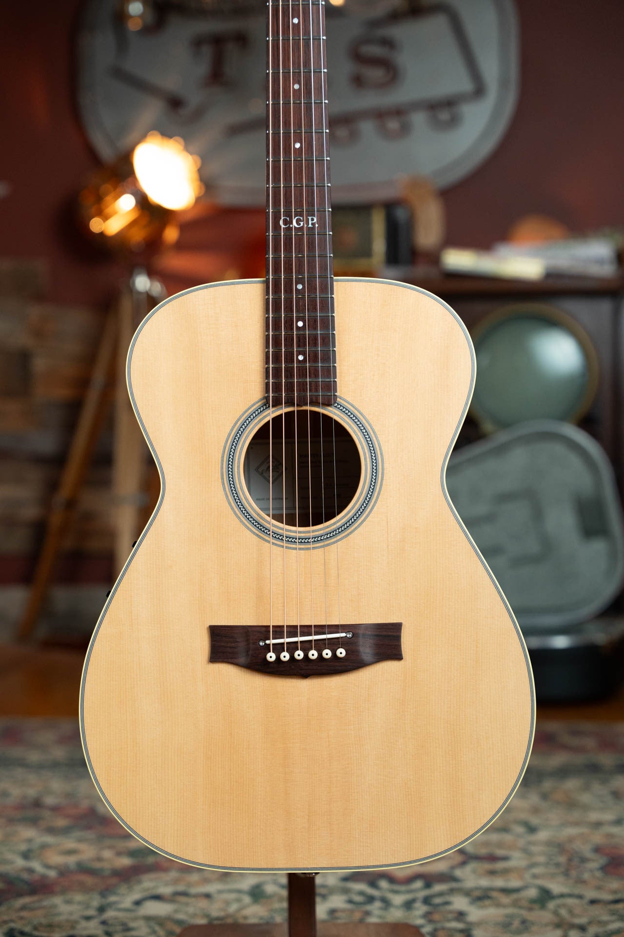 A Maton Guitars T.E. Personal Spruce/Queensland Maple Deep Body OO Guitar w/Electronics (#0085) with AP5 PRO pickup stands upright in a cozy, vintage-style room with patterned rug, wood accents, and blurred decor.