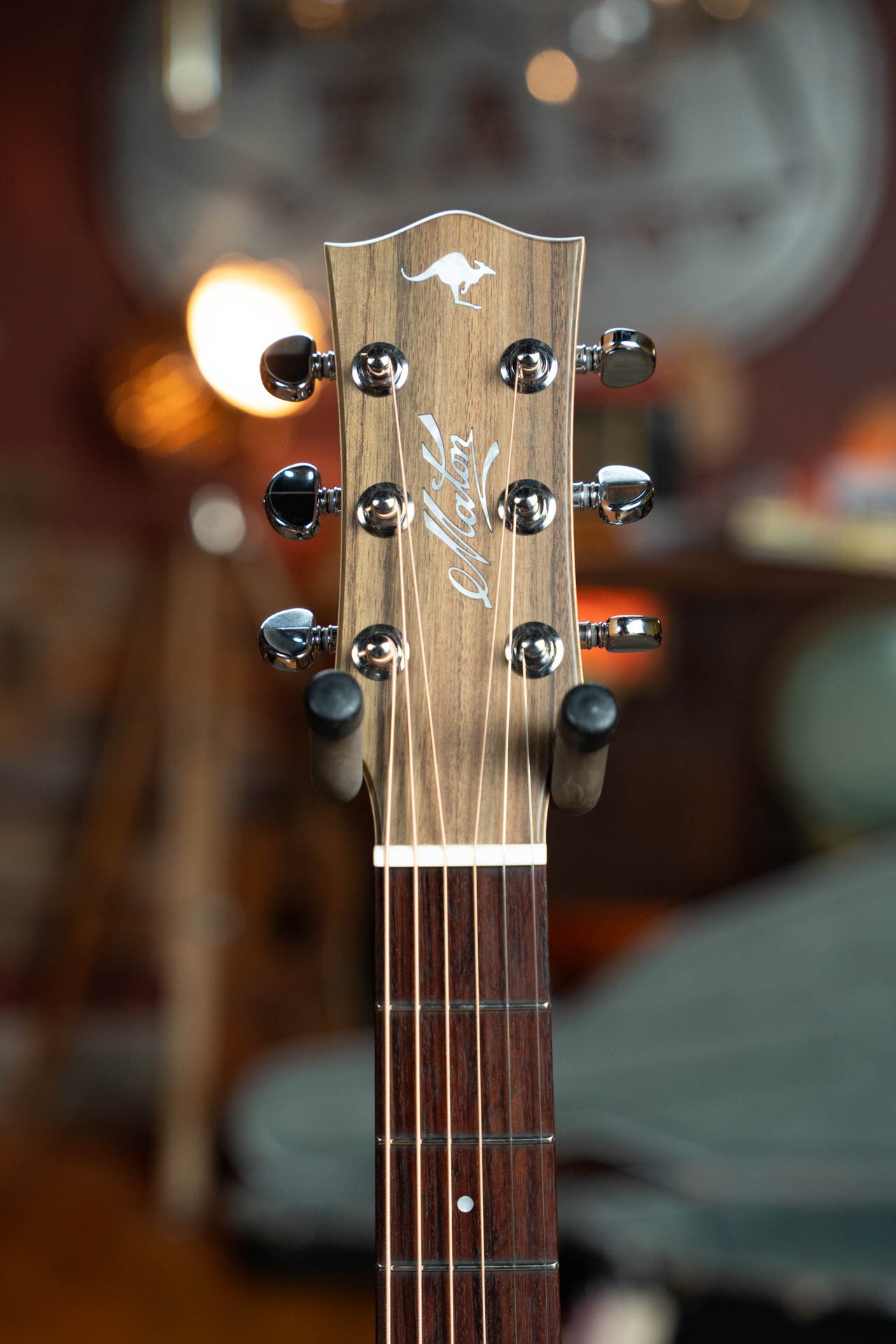 Close-up of the Maton Guitars T.E. Personal Spruce/Queensland Maple Deep Body OO Guitar headstock, featuring six tuners, Tommy Emmanuels signature, brand logo, and AP5 PRO electronics, set against a softly lit cozy background.