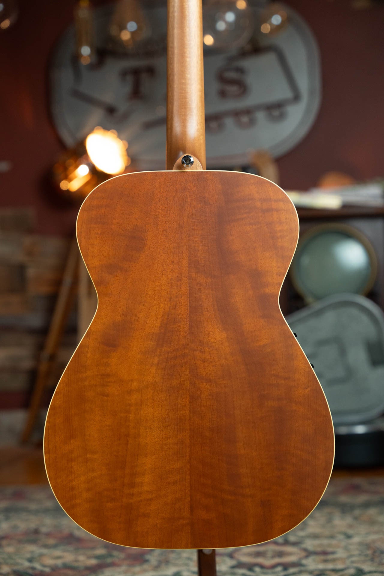 A close-up of the back of the Maton Guitars T.E. Personal Spruce/Queensland Maple Deep Body OO Guitar w/Electronics #0085, showcasing its smooth wood grain finish in a warmly lit, music-themed room.