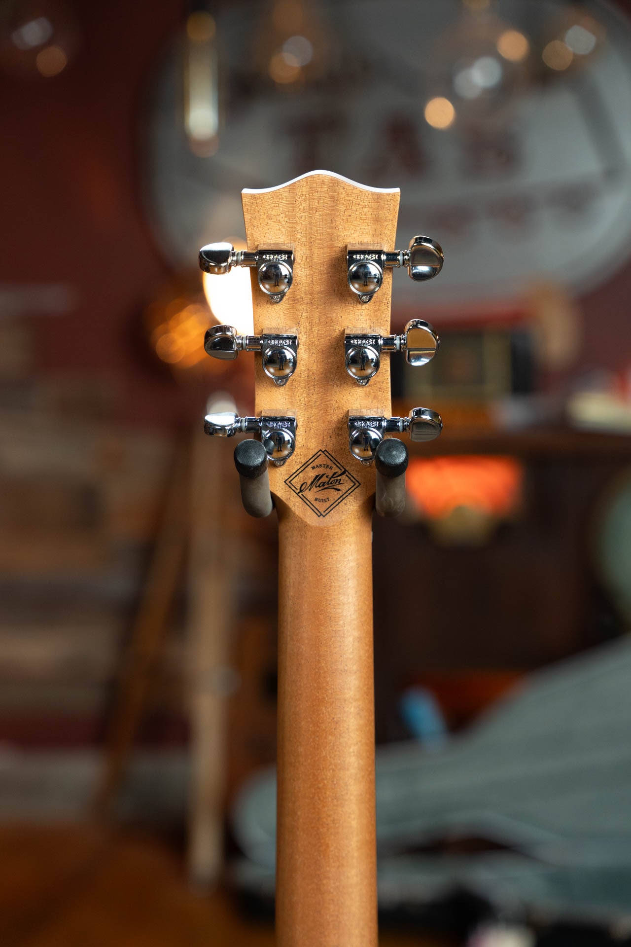 Close-up of the Maton Guitars T.E. Personal Spruce/Queensland Maple Deep Body OO Guitar #0085 headstock, highlighting six chrome tuners and logo, with a cozy, warmly lit room featuring wooden accents in the background.