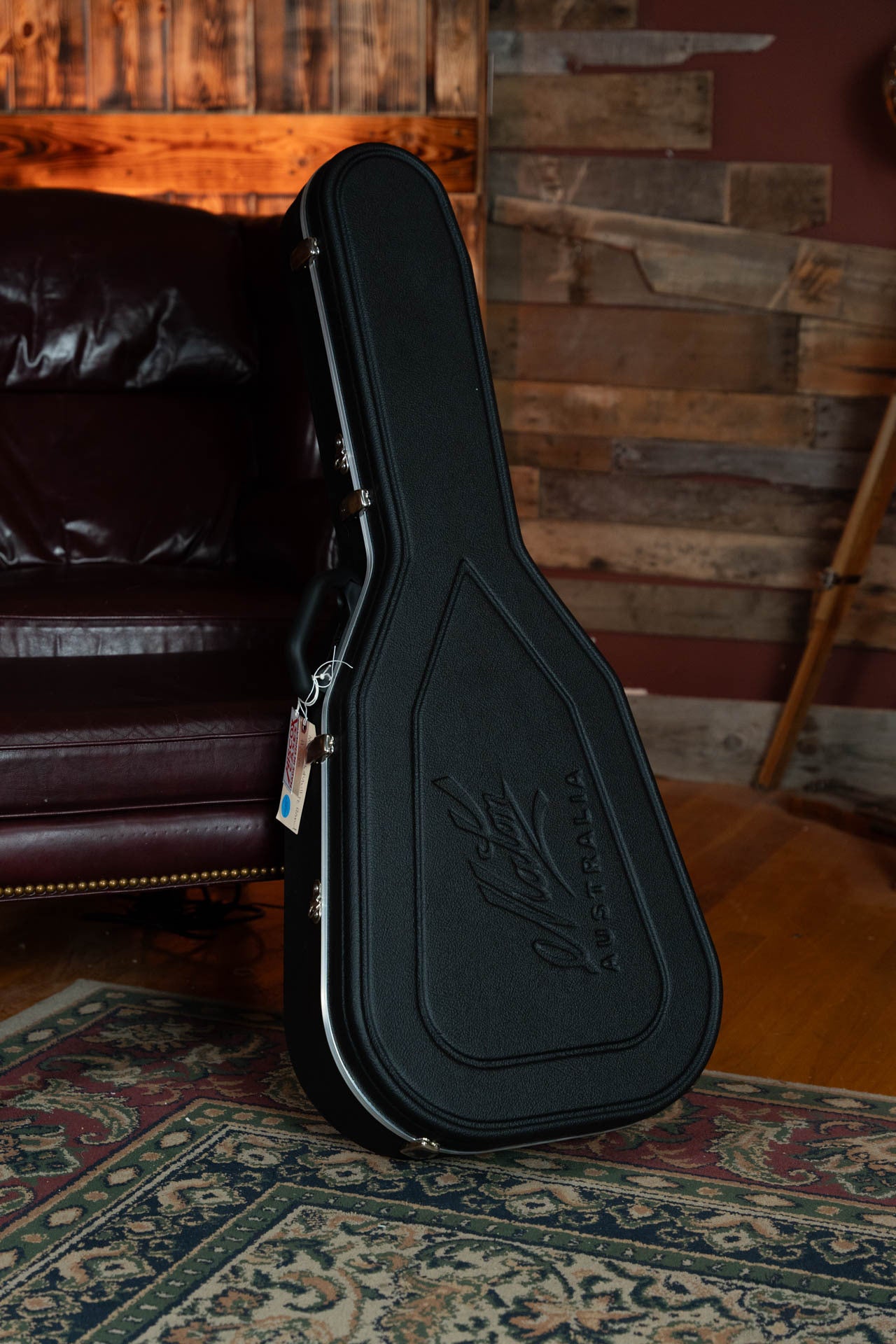 A black Maton Guitars case, embossed with Maton Australia, stands upright on a patterned rug before a leather couch and wooden paneled wall—ready to hold the Maton T.E. Personal Spruce/Queensland Maple Deep Body OO Guitar #0085.