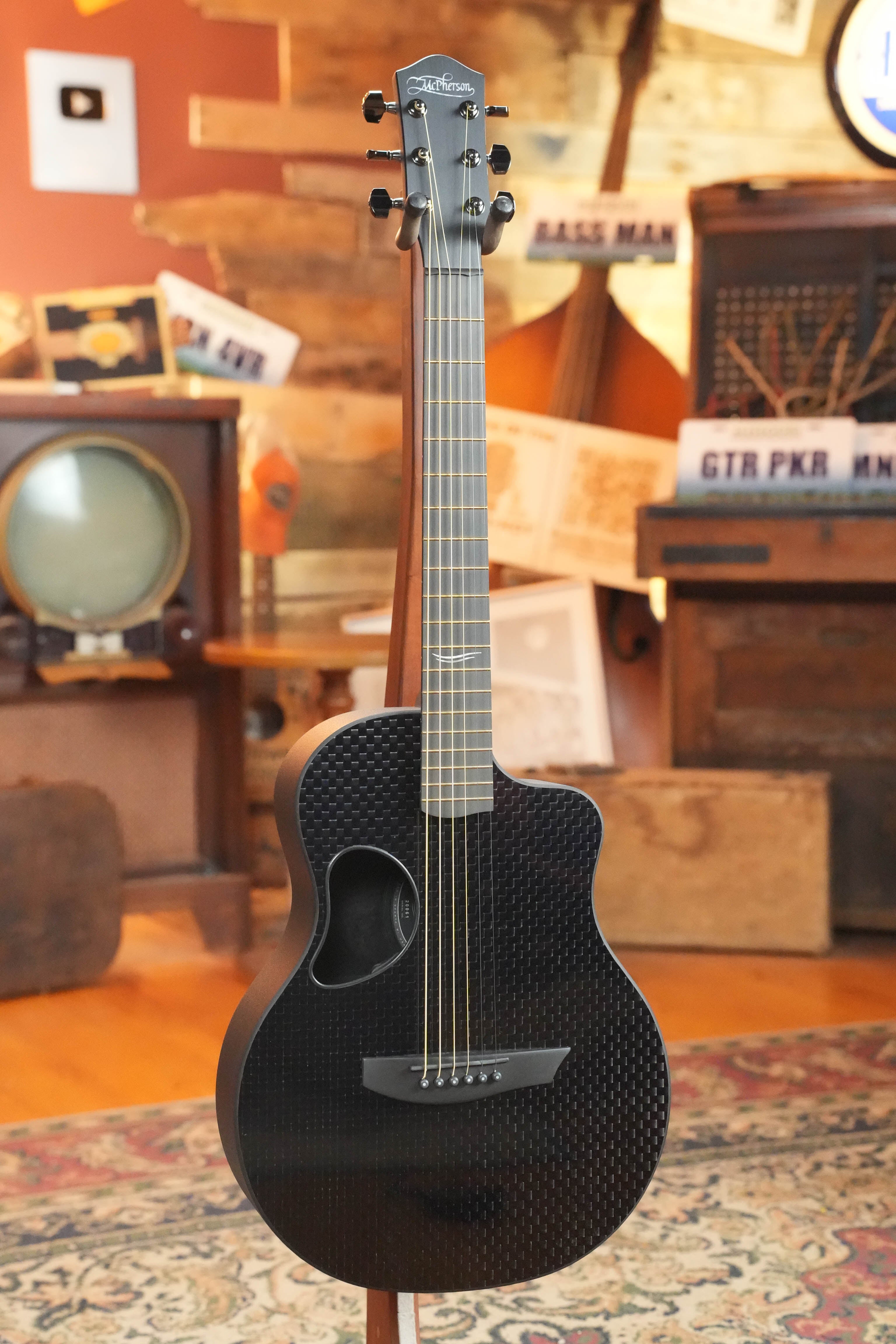 A McPherson Guitars Carbon Fiber Touring Series Blackout Basketweave Guitar w/L.R. Baggs Element Pickup (#0061) stands on a wooden floor in a cozy, vintage-themed room with an old TV, wooden furniture, and music signs in the background.