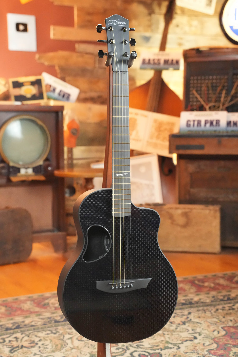 A McPherson Guitars Carbon Fiber Touring Series Blackout Basketweave Guitar w/L.R. Baggs Element Pickup (#0061) stands on a wooden floor in a cozy, vintage-themed room with an old TV, wooden furniture, and music signs in the background.