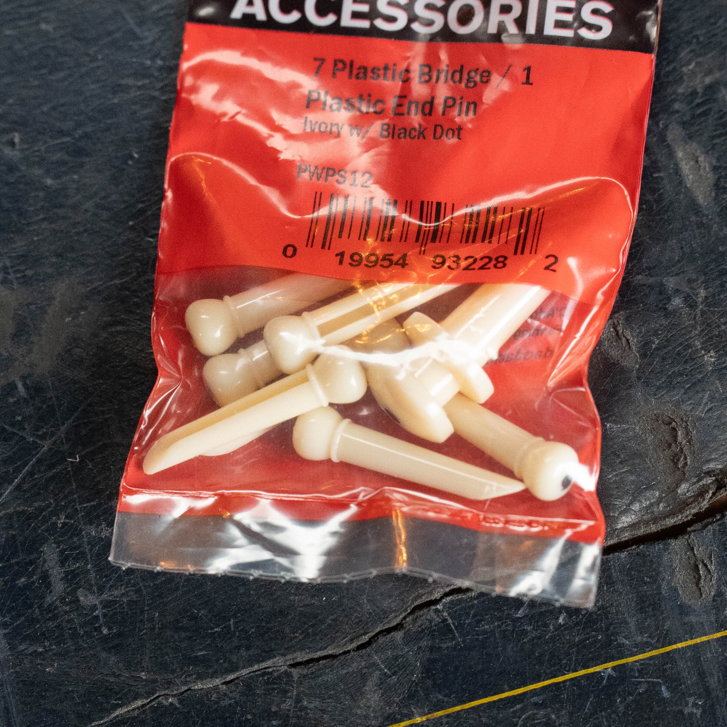 A package of D'Addario Injected Molded Bridge Pins with End Pin (PWPS12), Set of 7, Ivory with Ebony Dot, rests on a dark textured surface.