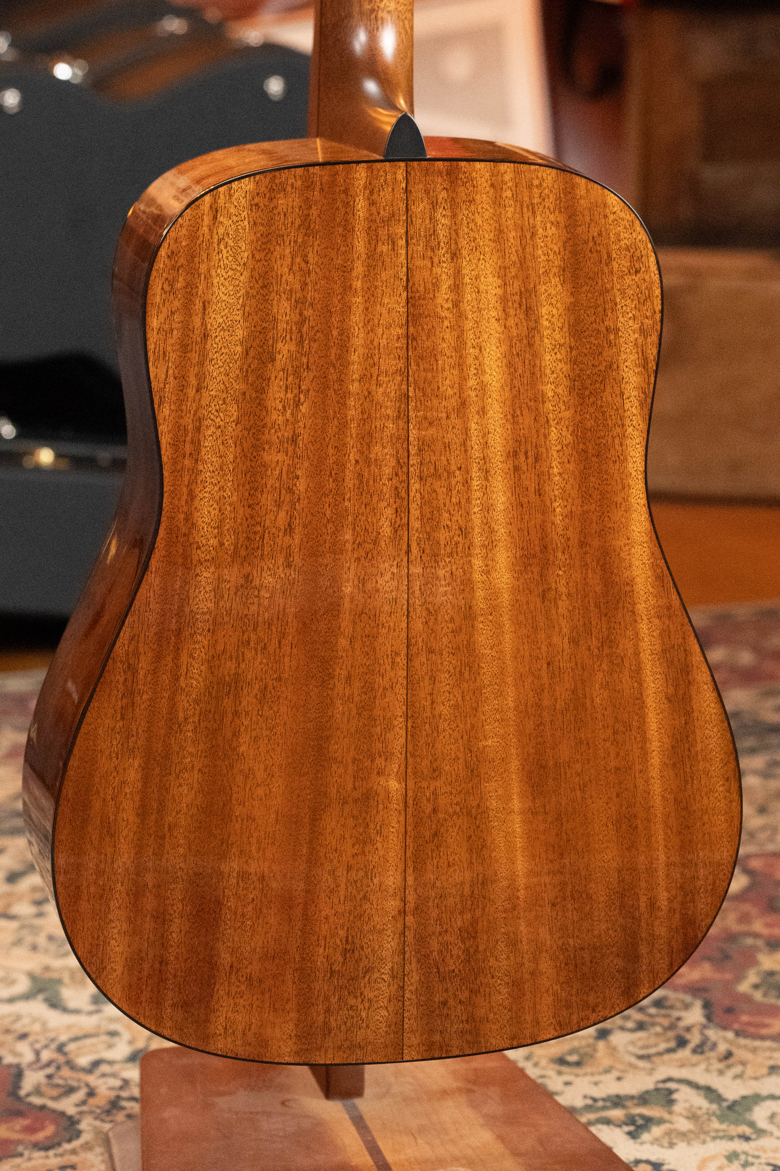 Close-up of the back of a Martin & Co. *New 2025 Model* D-18 1933 Gloss Ambertone Spruce/Mahogany Dreadnought Acoustic Guitar #6784, upright on a stand, highlighting its polished finish and wood grain with a patterned rug behind.