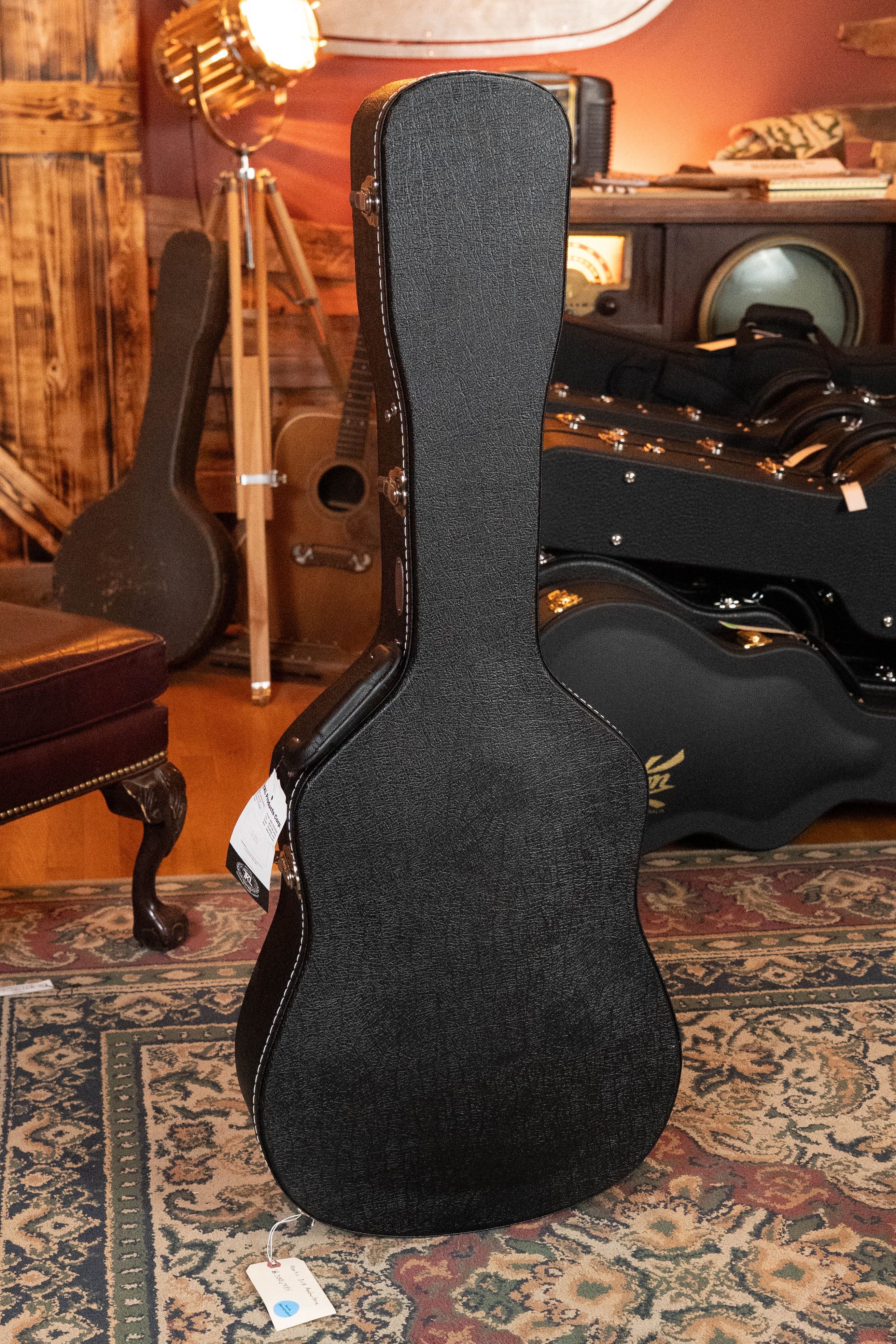 A black hard guitar case stands upright on a patterned rug in a vintage-themed room—ideal for storing the *New 2025 Model* Martin & Co. D-18 1933 Gloss Ambertone Spruce/Mahogany Dreadnought Acoustic Guitar #6784.