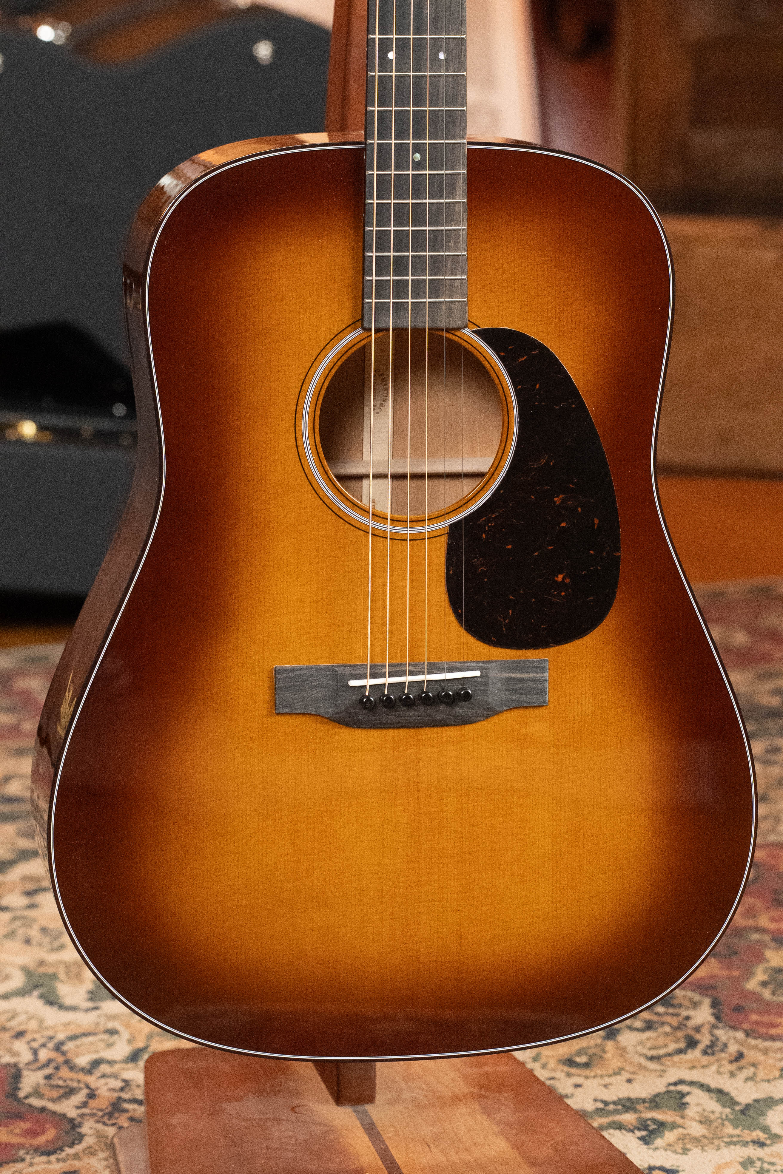 A close-up of the *New 2025 Model* Martin D-18 1933 Gloss Ambertone Spruce/Mahogany Dreadnought Acoustic Guitar #6784 by Martin & Co., upright on a stand over a patterned rug, shows off its sunburst finish and black pickguard.