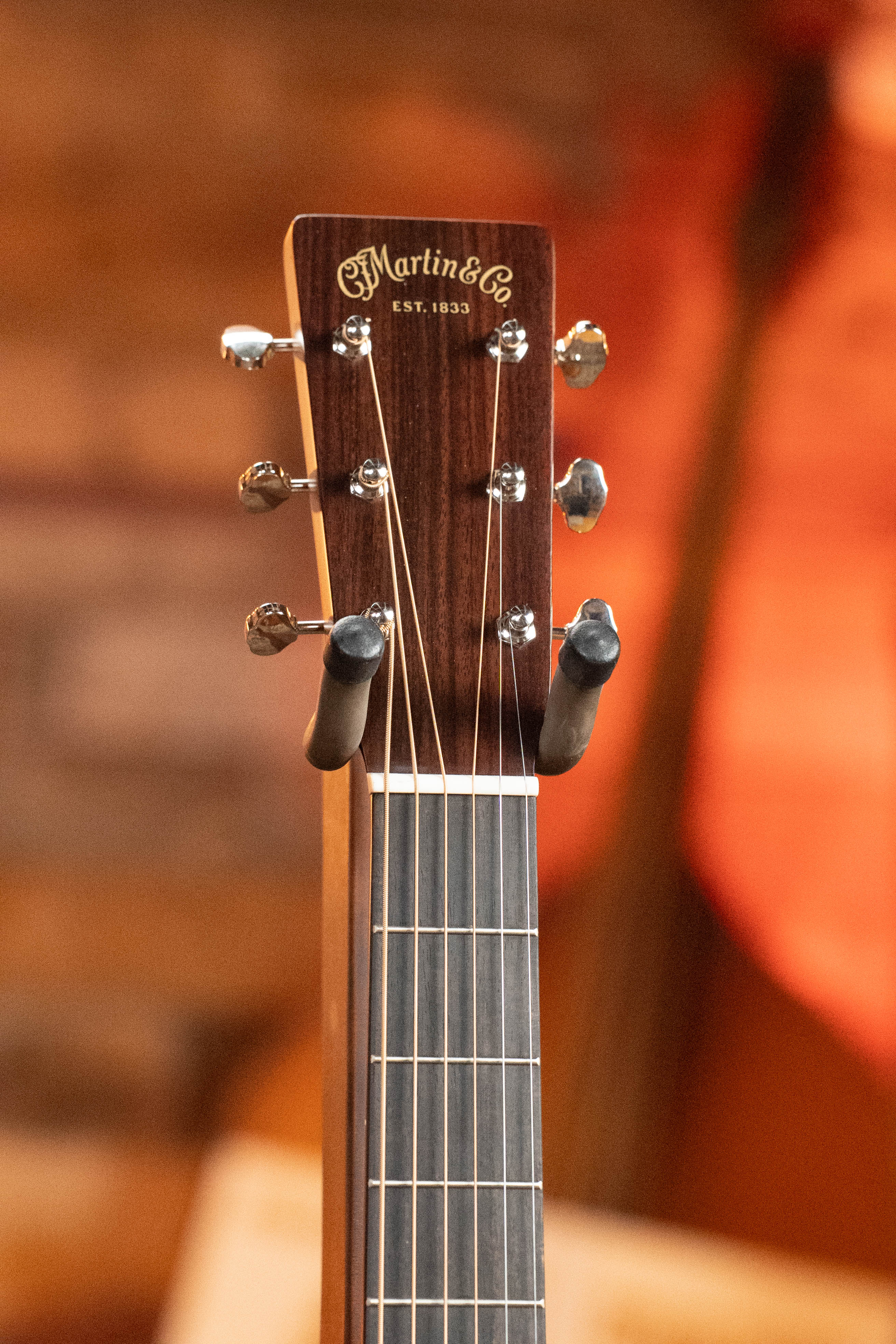 Close-up of a Martin & Co. New 2025 Model D-18 1933 Gloss Ambertone Spruce/Mahogany dreadnought acoustic guitar (#6784) headstock with visible tuning pegs and logo, resting on a stand against a warm, blurred background.