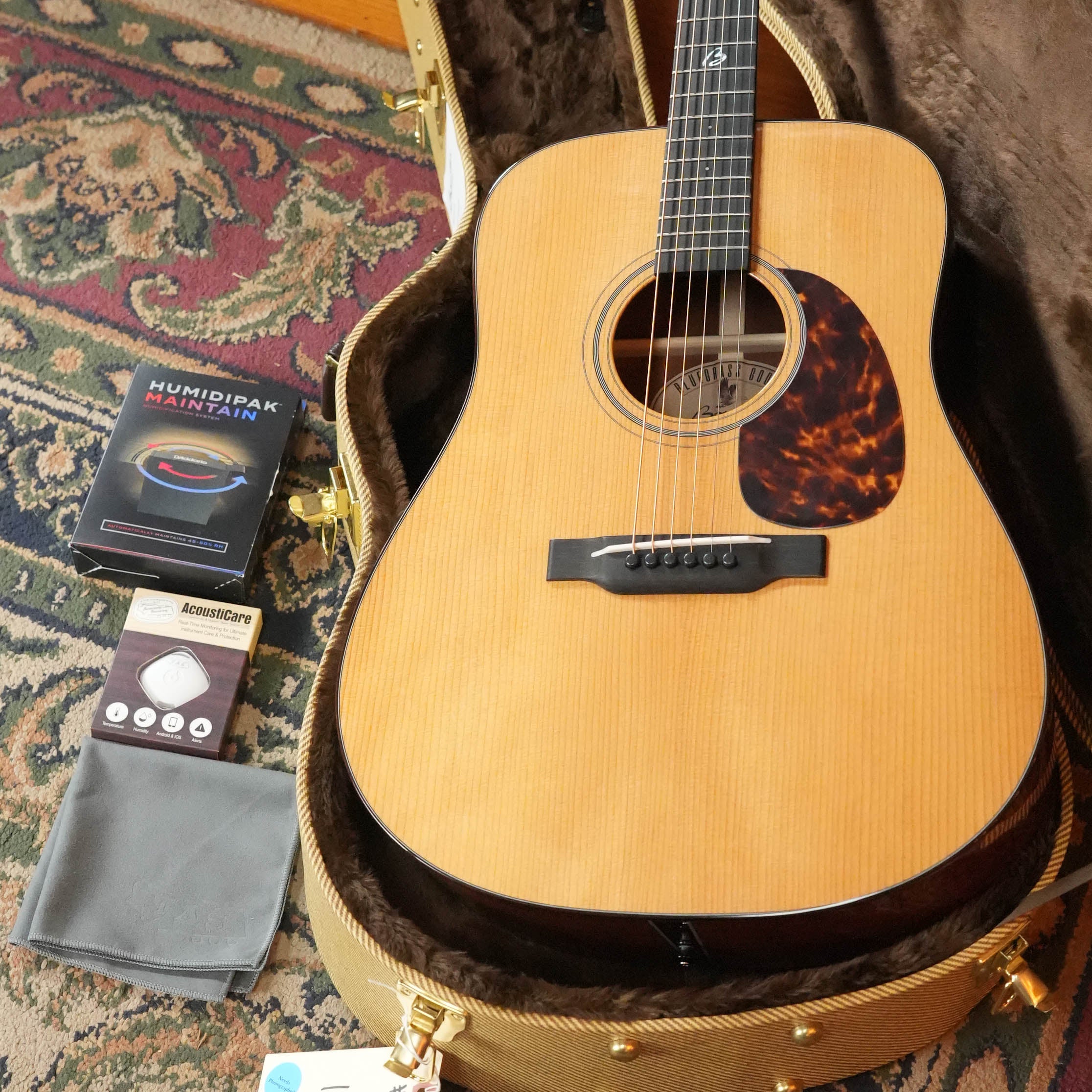 A *New 2026 Model* Boucher BG-42T-G Torrefied Adirondack/Torrefied Mahogany dreadnought #1082 by Boucher Guitars rests in its hardshell case, with Humidipak, AcousticCare product, and gray cleaning cloth on a patterned rug.