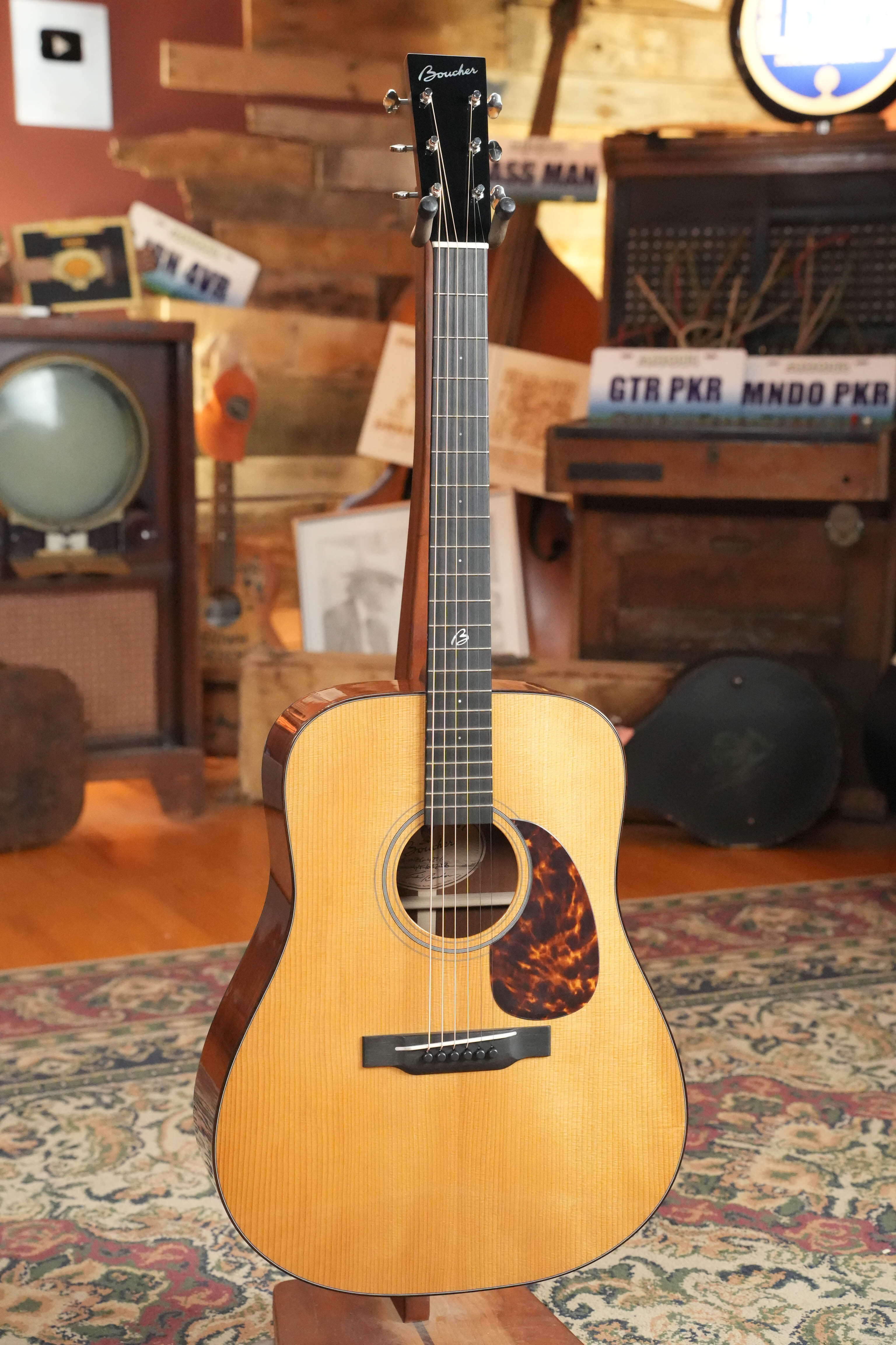 A *New 2026 Model* Boucher BG-42T-G Torrefied Adirondack/Torrefied Mahogany Bluegrass Goose Gold Pack Dreadnought Acoustic Guitar #1082 by Boucher Guitars stands on a colorful rug in a cozy, music-themed room with vintage decor and wooden walls.