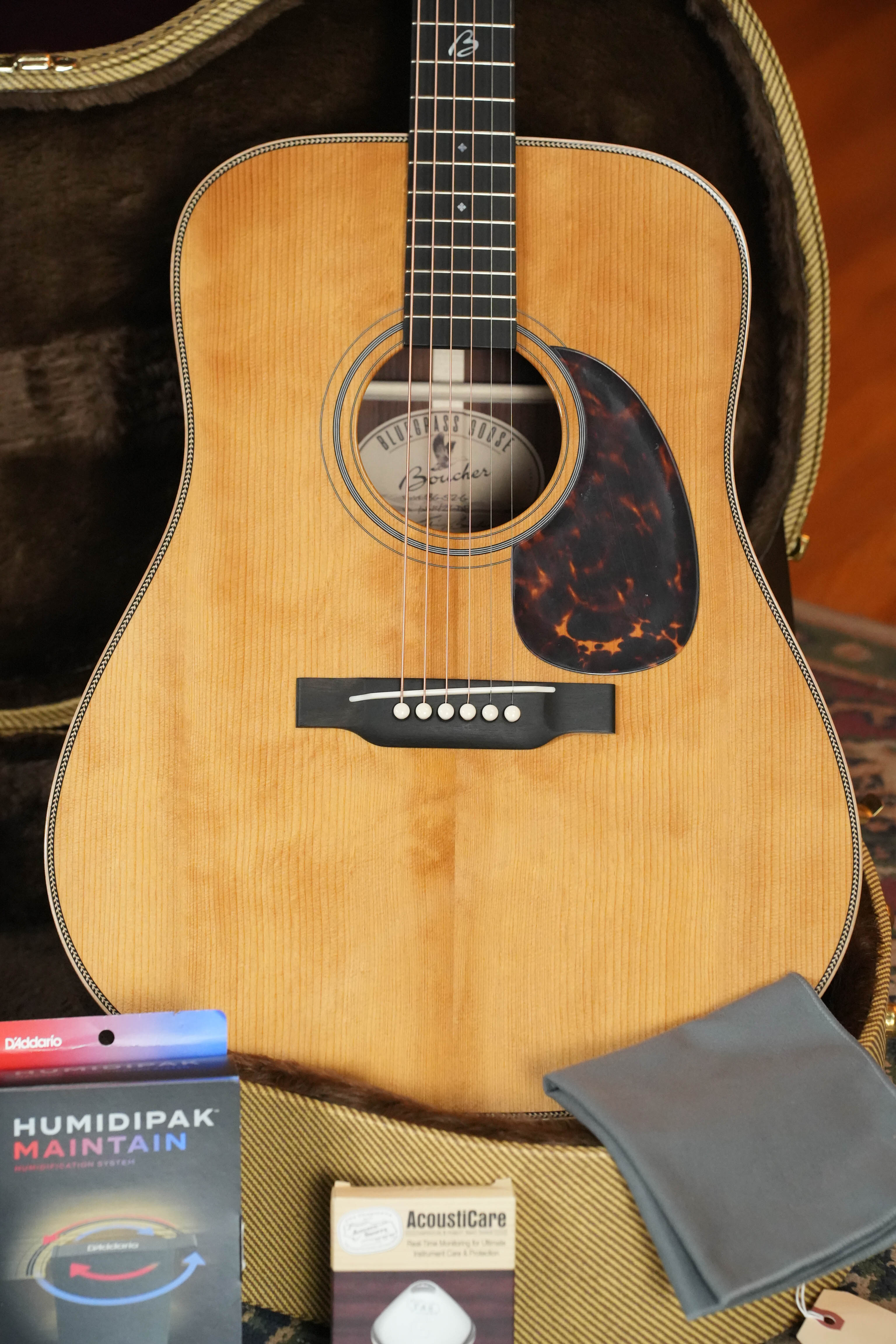 A *New 2026 Model* Boucher BG-52-G Torrefied Adirondack/Rosewood Bluegrass Goose Gold Pack Dreadnought Acoustic Guitar #1421 by Boucher Guitars rests in an open case, with a Humidipak, AcousticCare cleaner, and gray cloth neatly arranged in front.