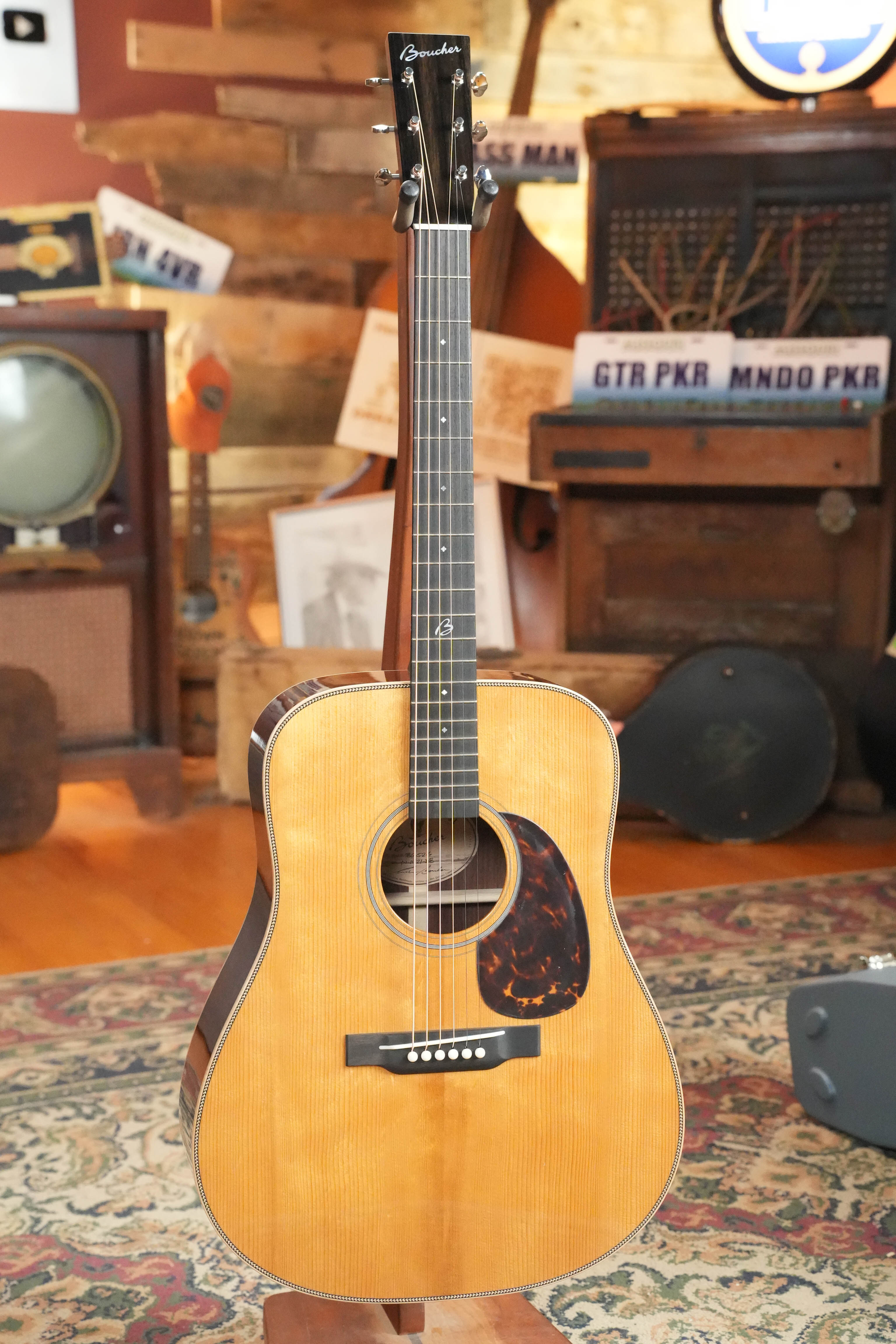 A *New 2026 Model* Boucher BG-52-G Torrefied Adirondack/Rosewood Bluegrass Goose Gold Pack dreadnought by Boucher Guitars, featuring a tortoiseshell pickguard, stands on a patterned rug in a vintage-inspired room with wooden décor.