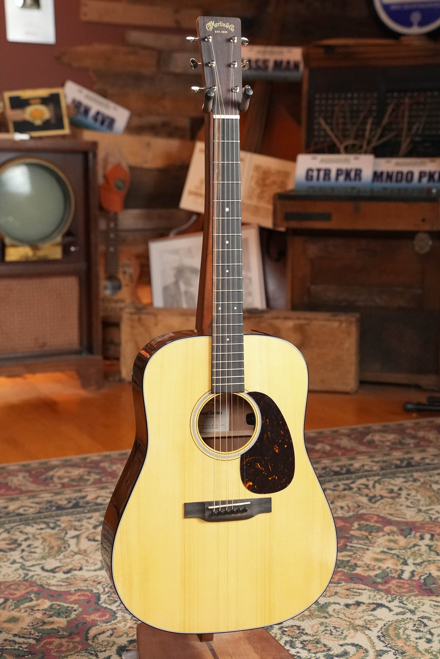 A *New 2026 Model* Martin D-12E Retro Road Series Spruce/Sapele Dreadnought Guitar by Martin & Co. (#1643) with a dark pickguard stands upright in a cozy vintage room with wood floors, retro decor, and old license plates visible.