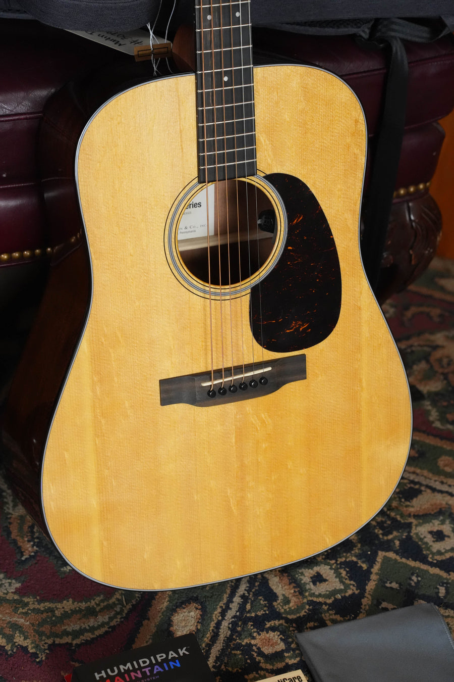 A close-up of the *New 2026 Model* Martin & Co. D-12E Retro Road Series Spruce/Sapele Dreadnought Guitar w/Martin Electronics #9387 stands upright on a patterned rug, displaying its light finish, sound hole, pickguard, and neck. Nearby guitar accessories are visible.