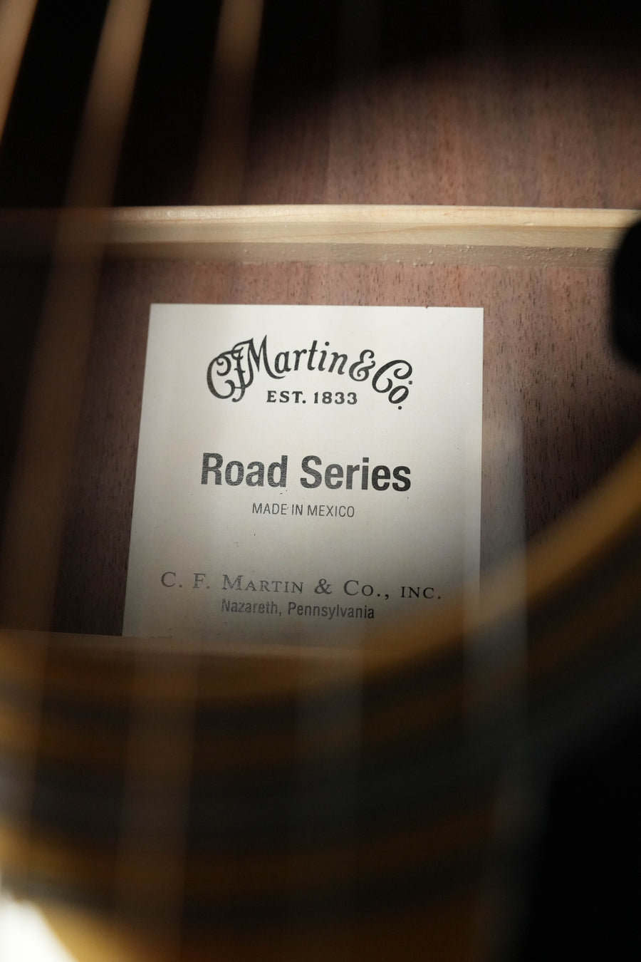 A close-up view through guitar strings reveals a label inside the Martin & Co. *New 2026 Model* D-13E Retro Road Series dreadnought, highlighting its quality craftsmanship and advanced Martin electronics.