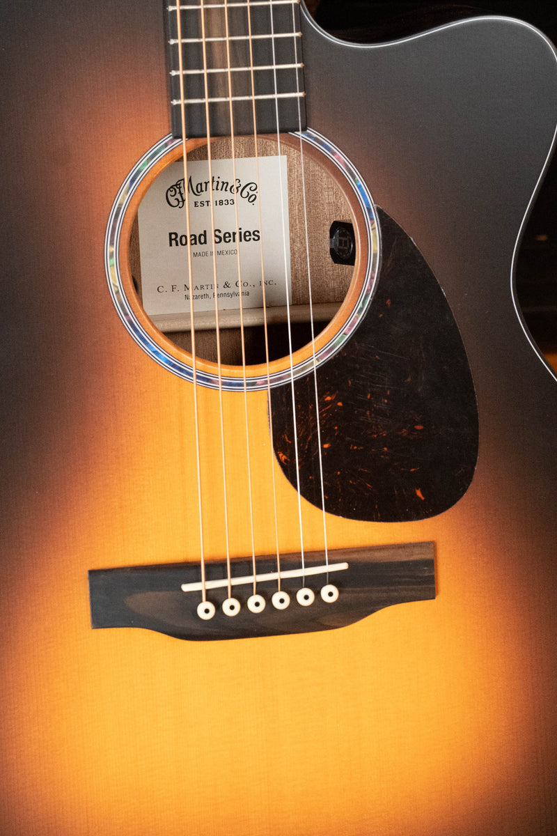Close-up of the *New 2026 Model* Martin OMC-10E Modern Burst Road Series Spruce/Sapele Orchestra Model Cutaway Guitar, showing strings, soundhole with Martin & Co. label, scalloped X-bracing for enhanced tone, bridge, and pickguard.