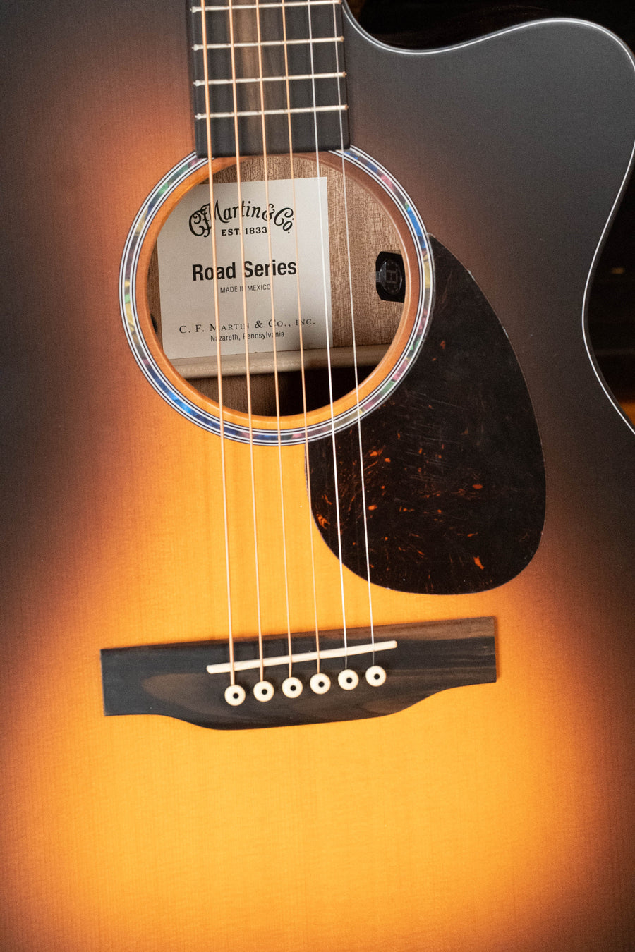 Close-up of the *New 2026 Model* Martin OMC-10E Modern Burst Road Series Spruce/Sapele Orchestra Model Cutaway Guitar, showing strings, soundhole with Martin & Co. label, scalloped X-bracing for enhanced tone, bridge, and pickguard.