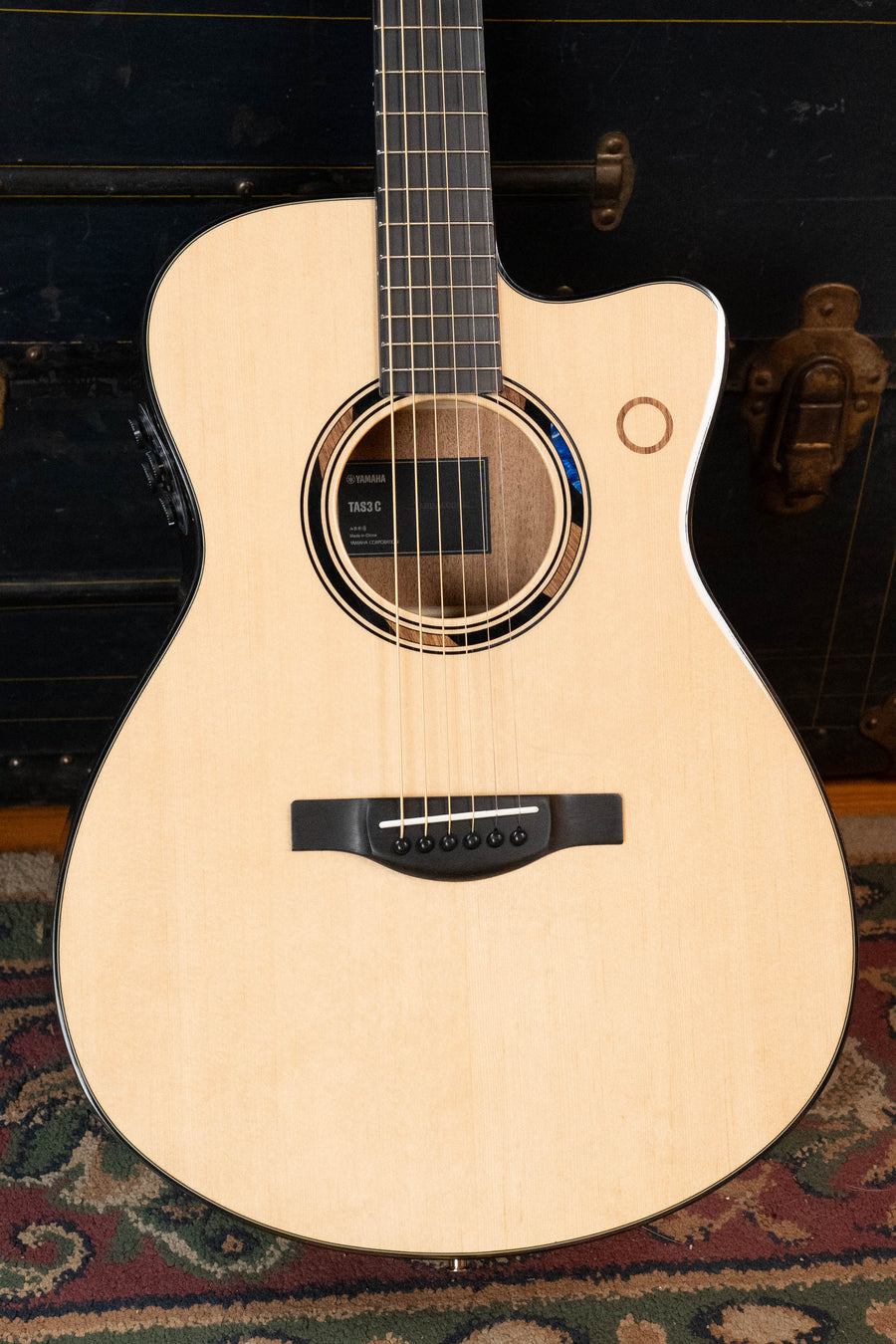 A close-up of the *New 2026 Model* Yamaha TAS3C-NT Sitka/Mahogany Concert Guitar with the latest TransAcoustic System, featuring a solid Sitka spruce top and black pickguard, leaning against a vintage trunk on a patterned rug.