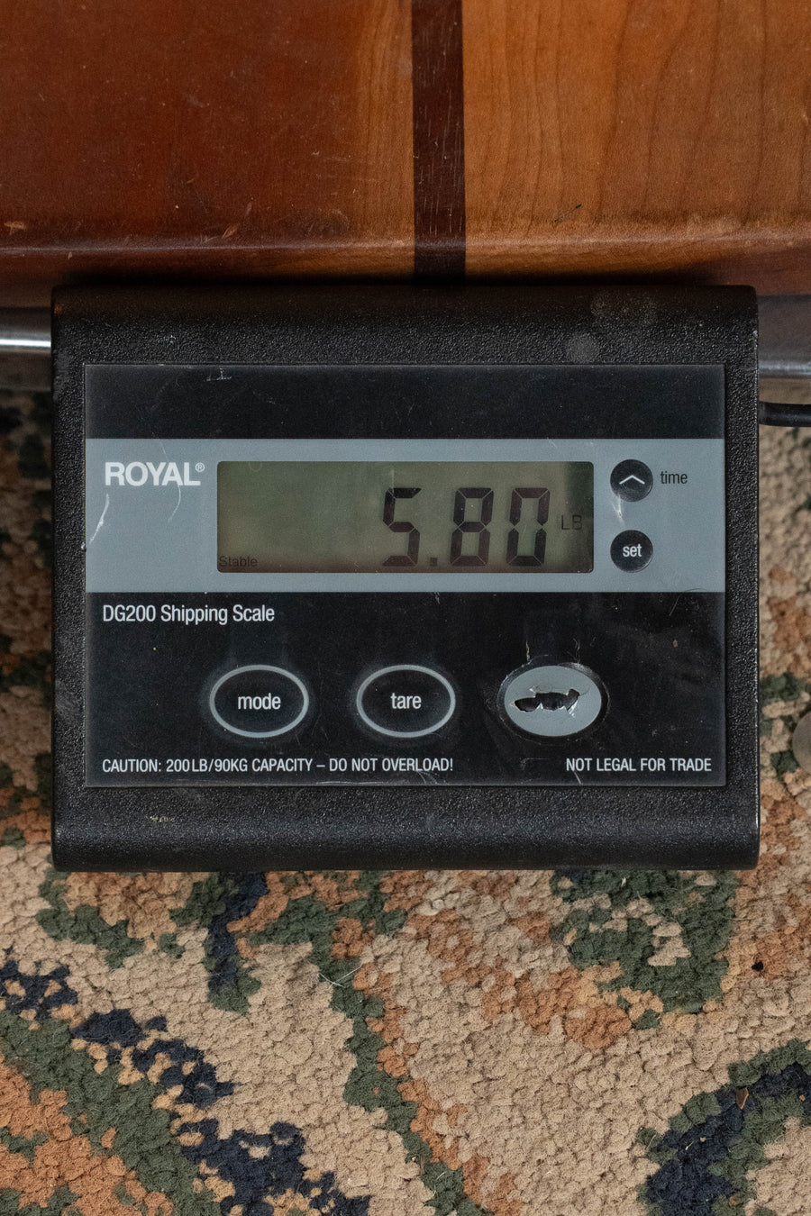 A digital shipping scale on a patterned carpet shows 5.80 pounds. The ROYAL DG200 Shipping Scale, with mode, tare, and settings buttons, is ideal for weighing items like the *New 2026 Model* Yamaha TAS3C-NT Sitka/Mahogany Concert Guitar.
