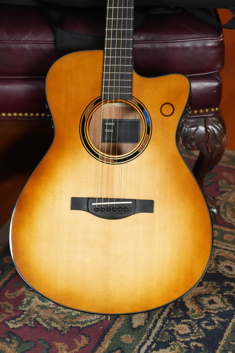 A close-up of a Yamaha TAS3C-SBD Sandburst Concert Guitar (*New 2026 Model*) featuring Sitka/Mahogany, with the newest TransAcoustic system, resting against a maroon chair with brass studs on a patterned carpet.