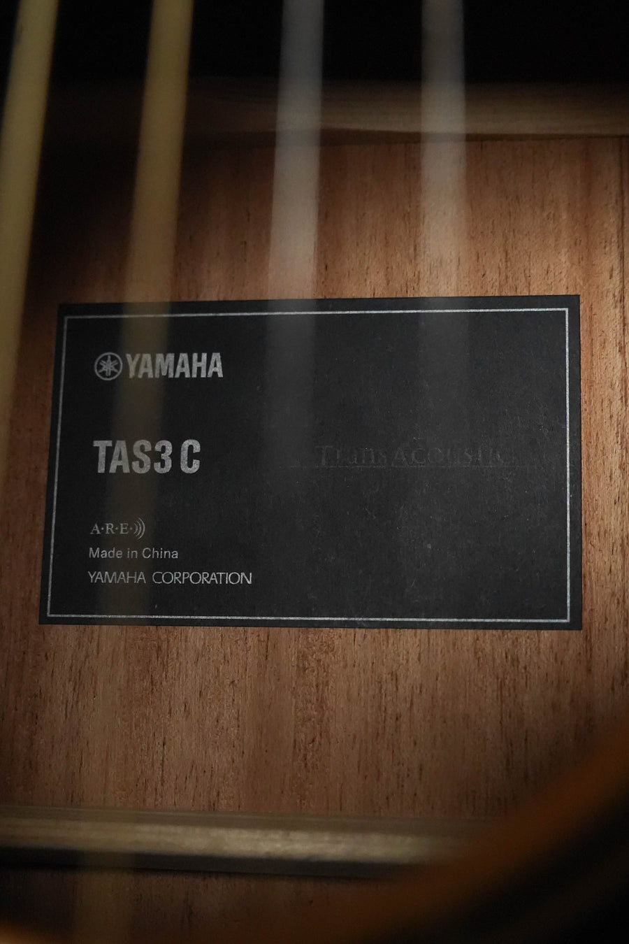 Close-up of a black label inside the Yamaha TAS3C-SBD Sitka/Mahogany Sandburst Concert Guitar, showing model #0165 and Yamaha Corporation, against the solid Sitka spruce top. Features the NEW 2026 model with the latest TransAcoustic system.