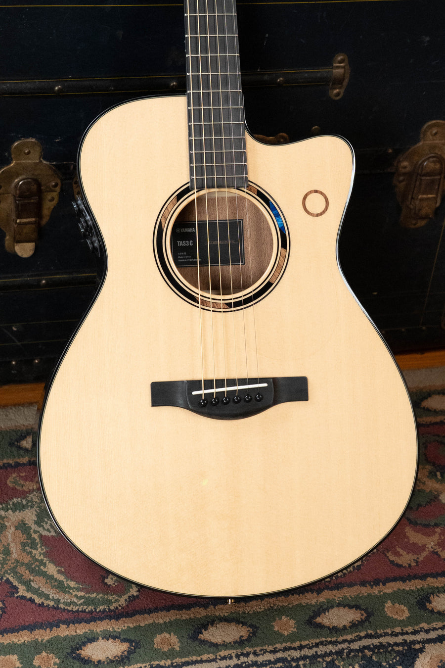 A *New 2026 Model* Yamaha TAS3C-NT Sitka/Mahogany Natural Concert Guitar with the latest TransAcoustic System rests on a patterned rug before a dark chest with brass accents; its cutaway body and soundhole rosette are visible.