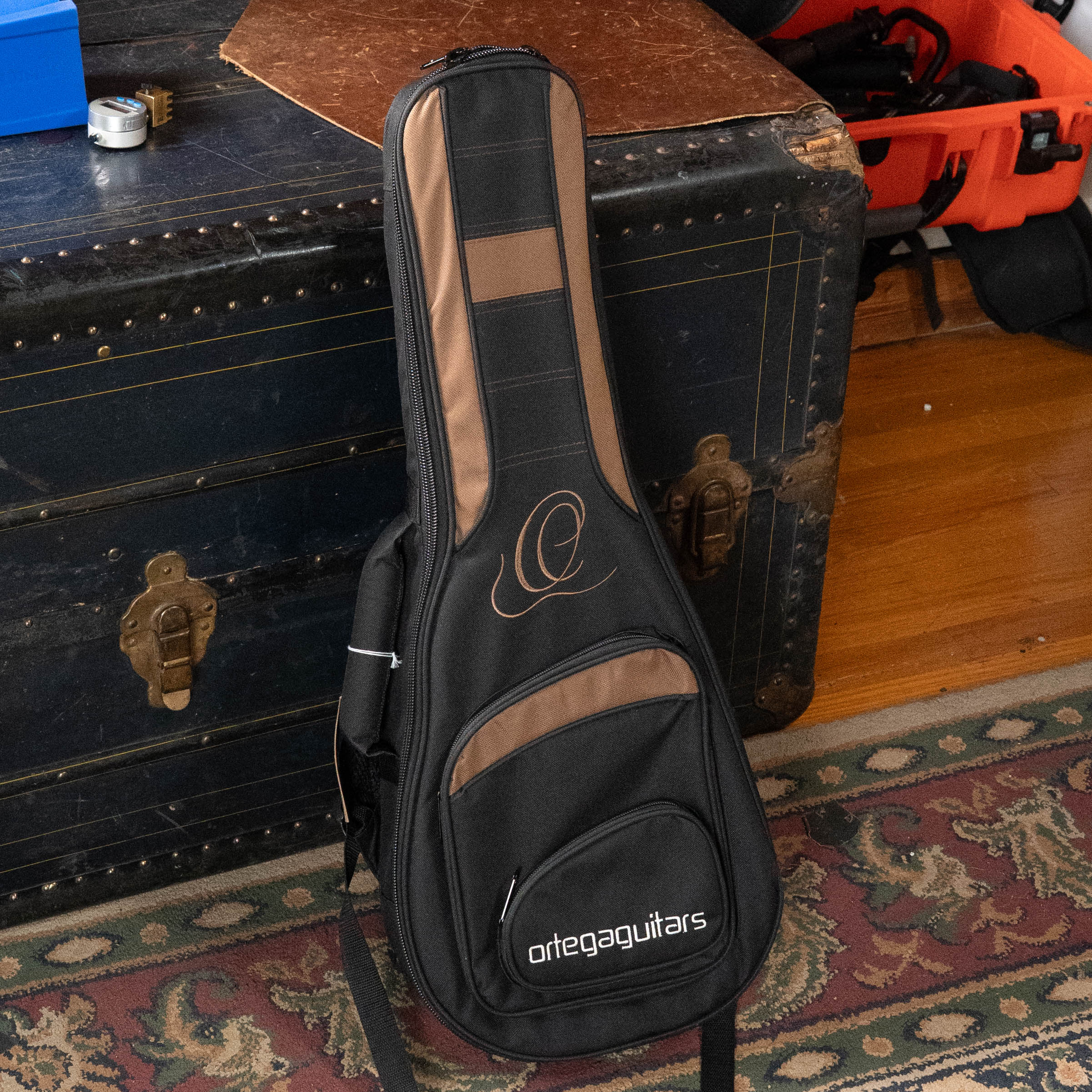 The Ortega ONB-MA Standard Size Mandolin Bag in brown with black by Ortega leans against a vintage trunk on a patterned rug, with toolboxes and assorted items in the background.