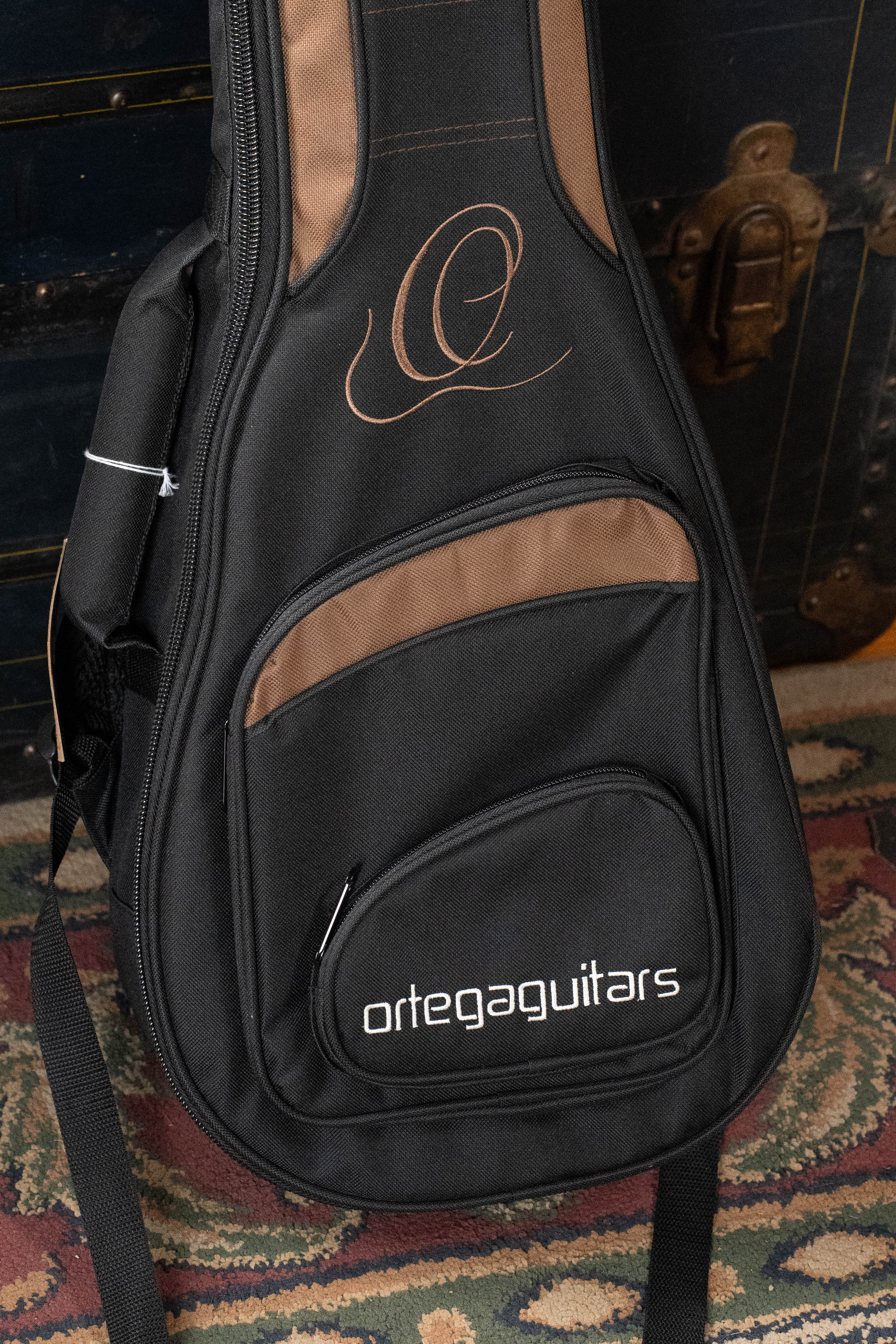 An Ortega ONB-MA Standard Size Mandolin Bag in brown with black from Ortega rests on a patterned rug, propped against a dark vintage trunk with metal hardware.