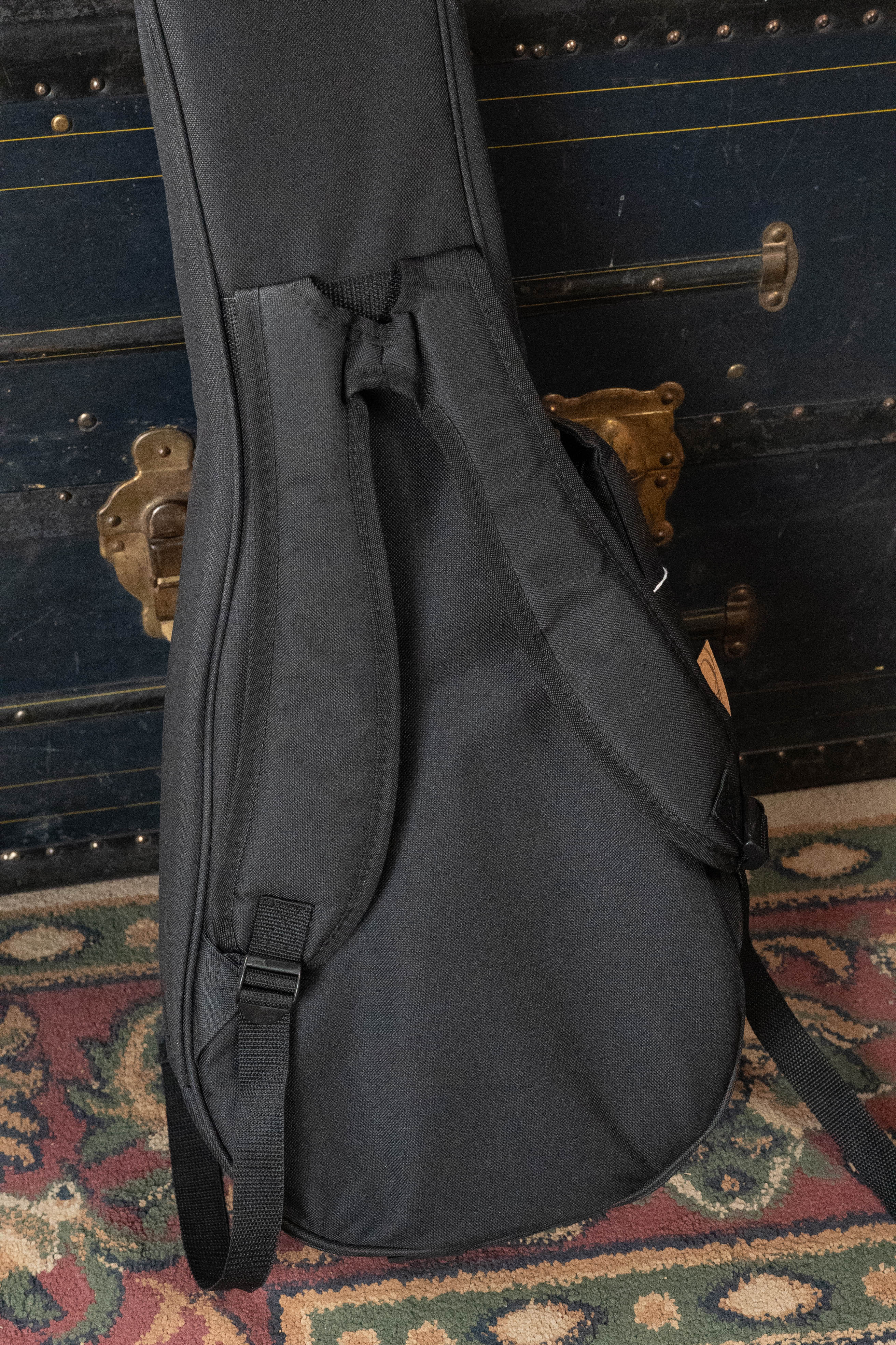 An Ortega ONB-MA Standard Size Mandolin Bag in brown with black stands upright on a colorful patterned rug, positioned in front of an old dark trunk with brass hardware.