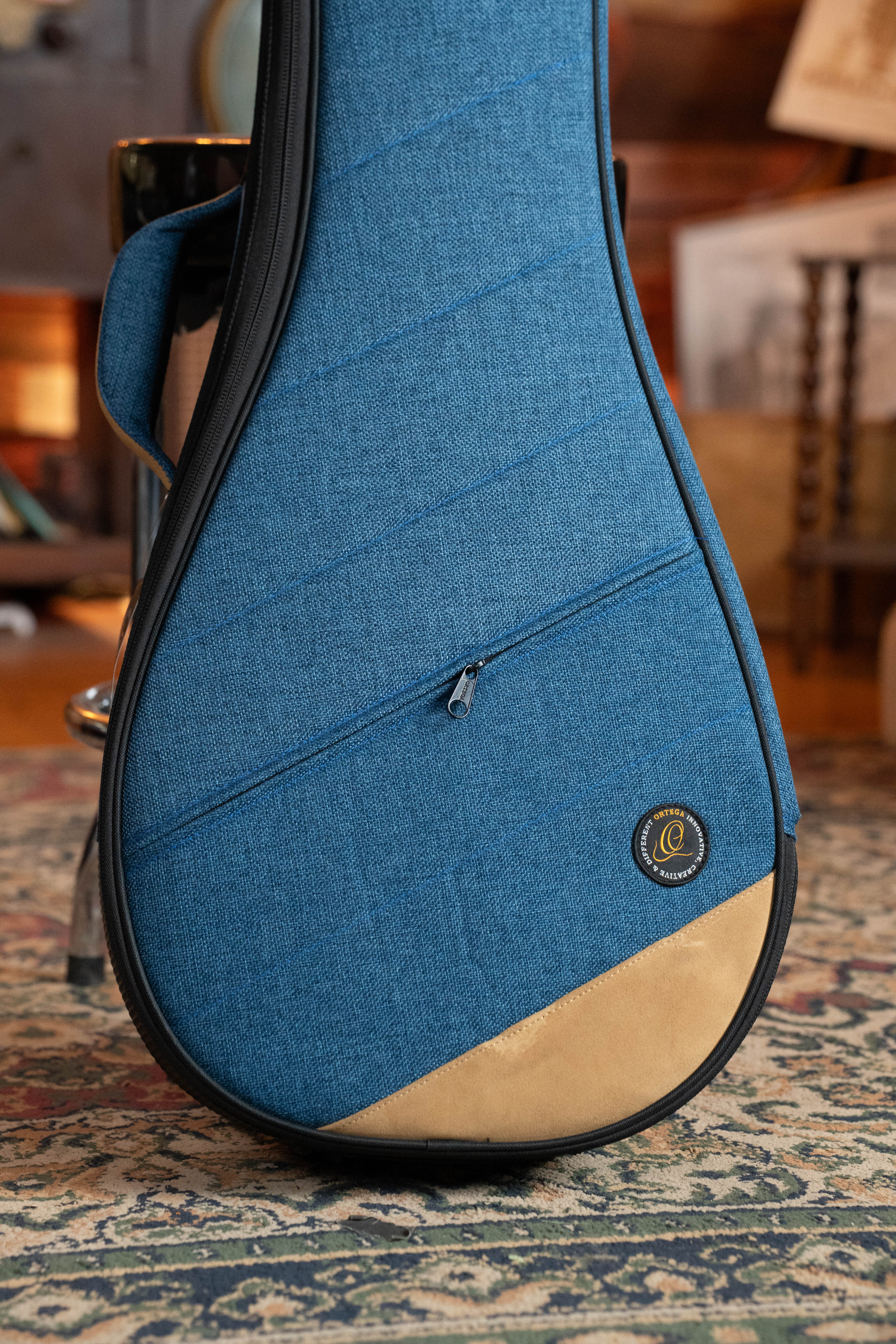 The Ortega OSOCABJ-OC Banjo Soft Case by Ortega, in ocean blue with a teardrop shape, tan accent, and small zippered pocket, stands upright on a patterned rug in a cozy, warmly lit room.