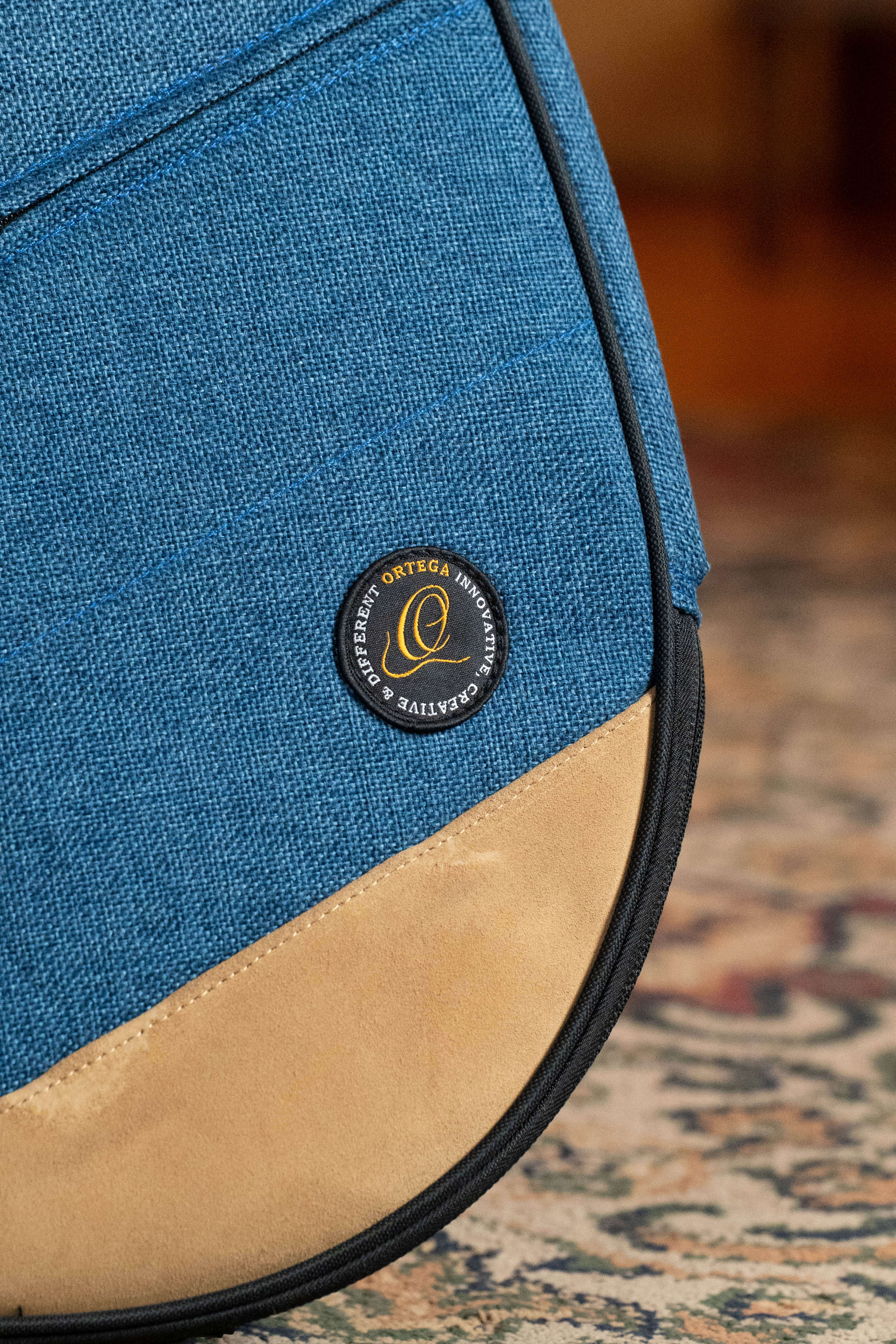 A close-up of the Ortega OSOCABJ-OC Banjo Soft Case in ocean blue, featuring a tan suede bottom and a round black-and-gold Ortega label. The case is set against a patterned carpet background.