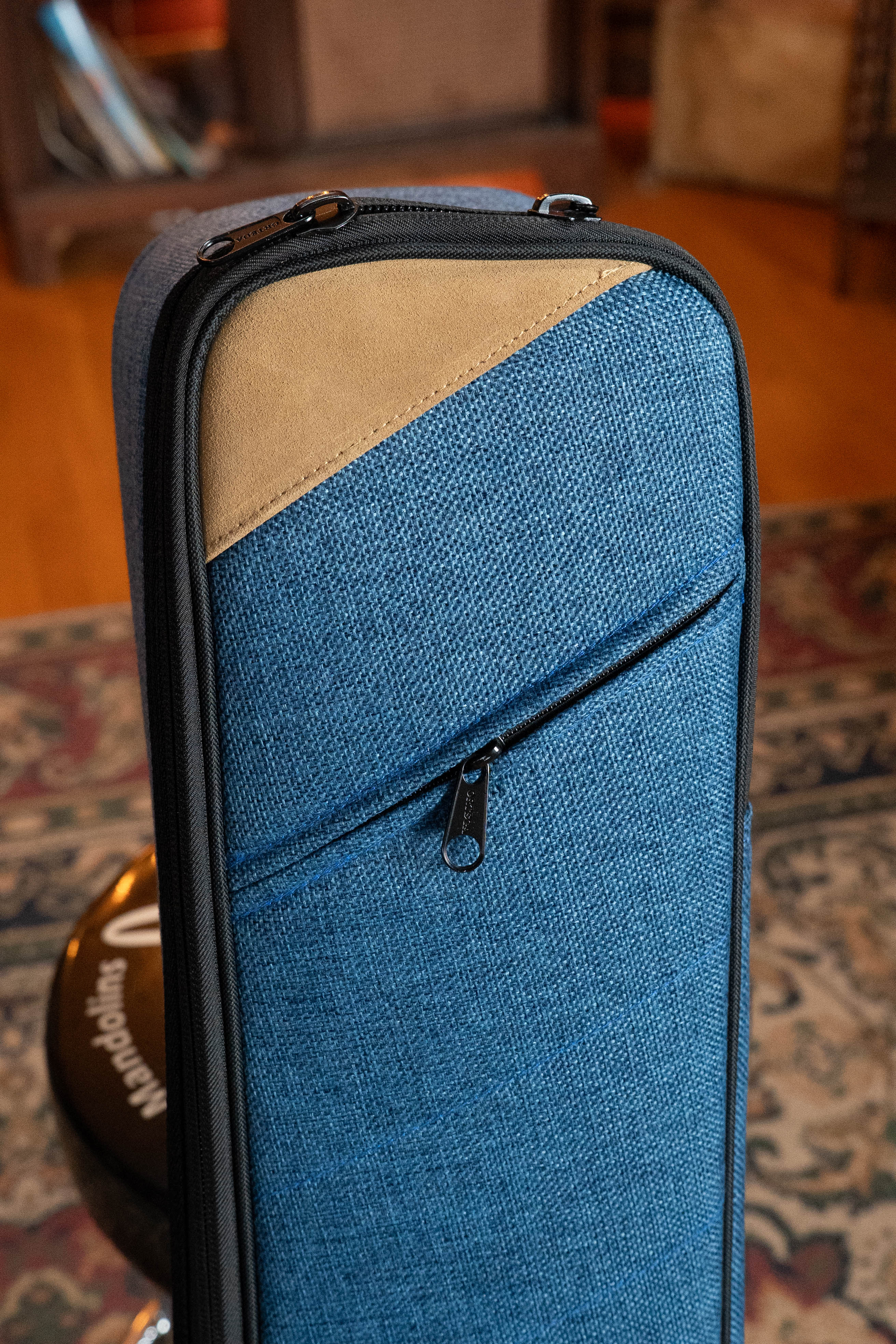 A close-up of the Ortega OSOCABJ-OC Banjo Soft Case in ocean blue with a tan accent and zippered pocket, shown on a patterned rug with bookshelves behind it.