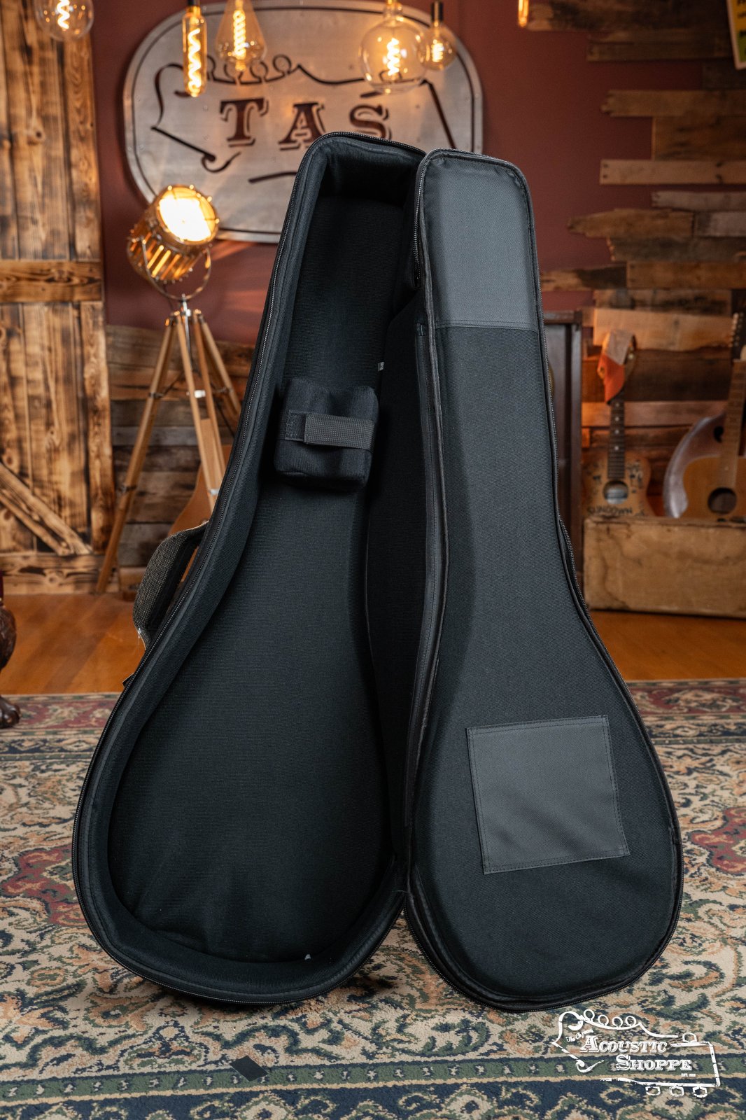 The Ortega OSOCABJ-MO Banjo Soft Case in Mocca by Ortega stands open on a patterned rug in a rustic room with wooden walls, guitars, and vintage lights in the background.
